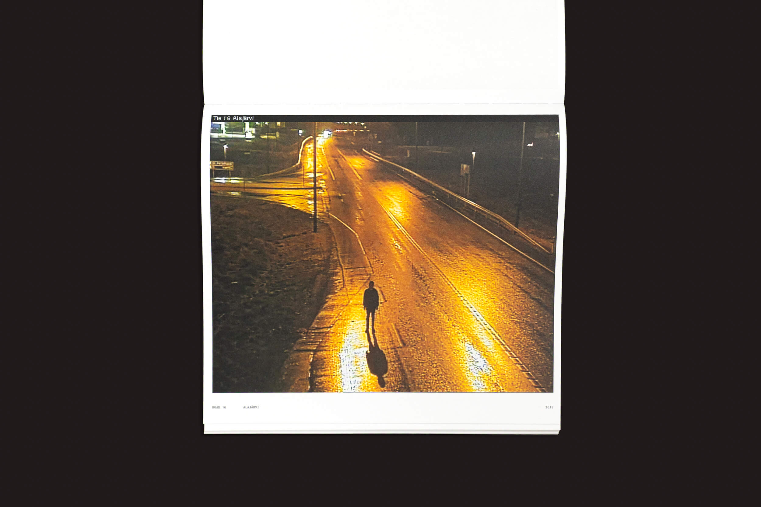 Uma foto capturando a solidão, com uma pessoa caminhando sozinha em uma rua molhada à noite, reflete o estilo de Tatu Gustafsson em "I on the Road / Weather Camera Self-Portraits, 2012-2021" da Fw:Books. As luzes da rua lançam um tom dourado quente, criando uma atmosfera pacífica e reflexiva.