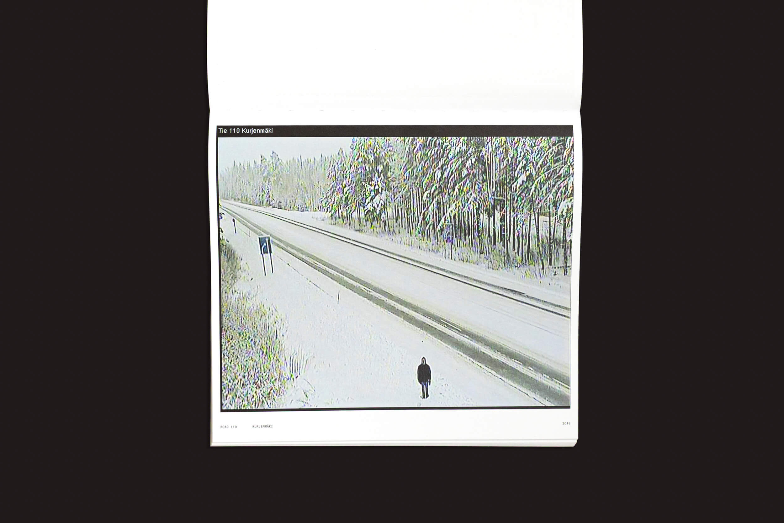 Uma figura solitária caminha por uma estrada coberta de neve, envolta por árvores cobertas de neve, em uma cena evocativa semelhante à fotografia alternativa, apresentada em "Tatu Gustafsson: I on the Road / Weather Camera Self-Portraits, 2012-2021" da Fw:Books.