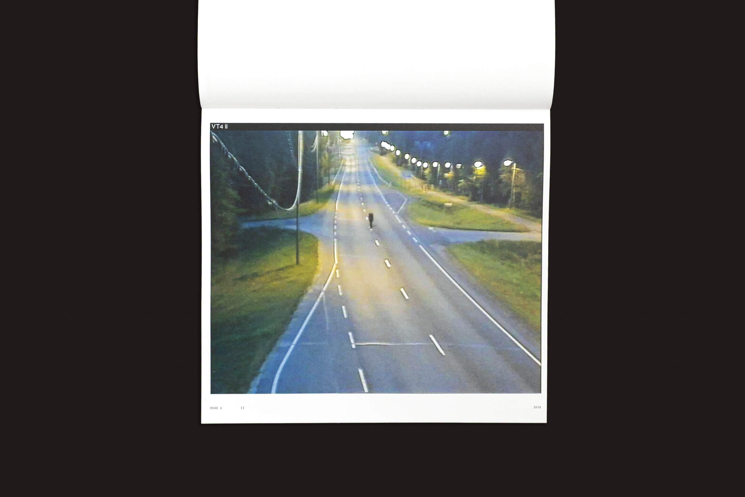 Um livro aberto revela uma foto de rua ao anoitecer de "Tatu Gustafsson: I on the Road / Weather Camera Self-Portraits, 2012-2021" de Fw:Books. A estrada iluminada apresenta um ciclista pedalando em direção à câmera, em um cenário escuro e arborizado.