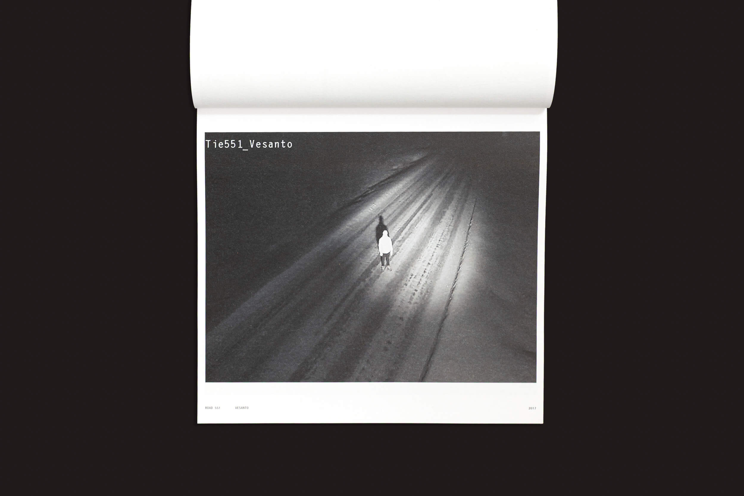Uma foto em preto e branco de "Tatu Gustafsson: I on the Road / Weather Camera Self-Portraits, 2012-2021" da Fw:Books mostra uma figura solitária em uma estrada com neve à noite, iluminada por uma luz. O canto superior esquerdo apresenta "TIE551, Vesanto", adicionando um toque enigmático que lembra fotografia alternativa.