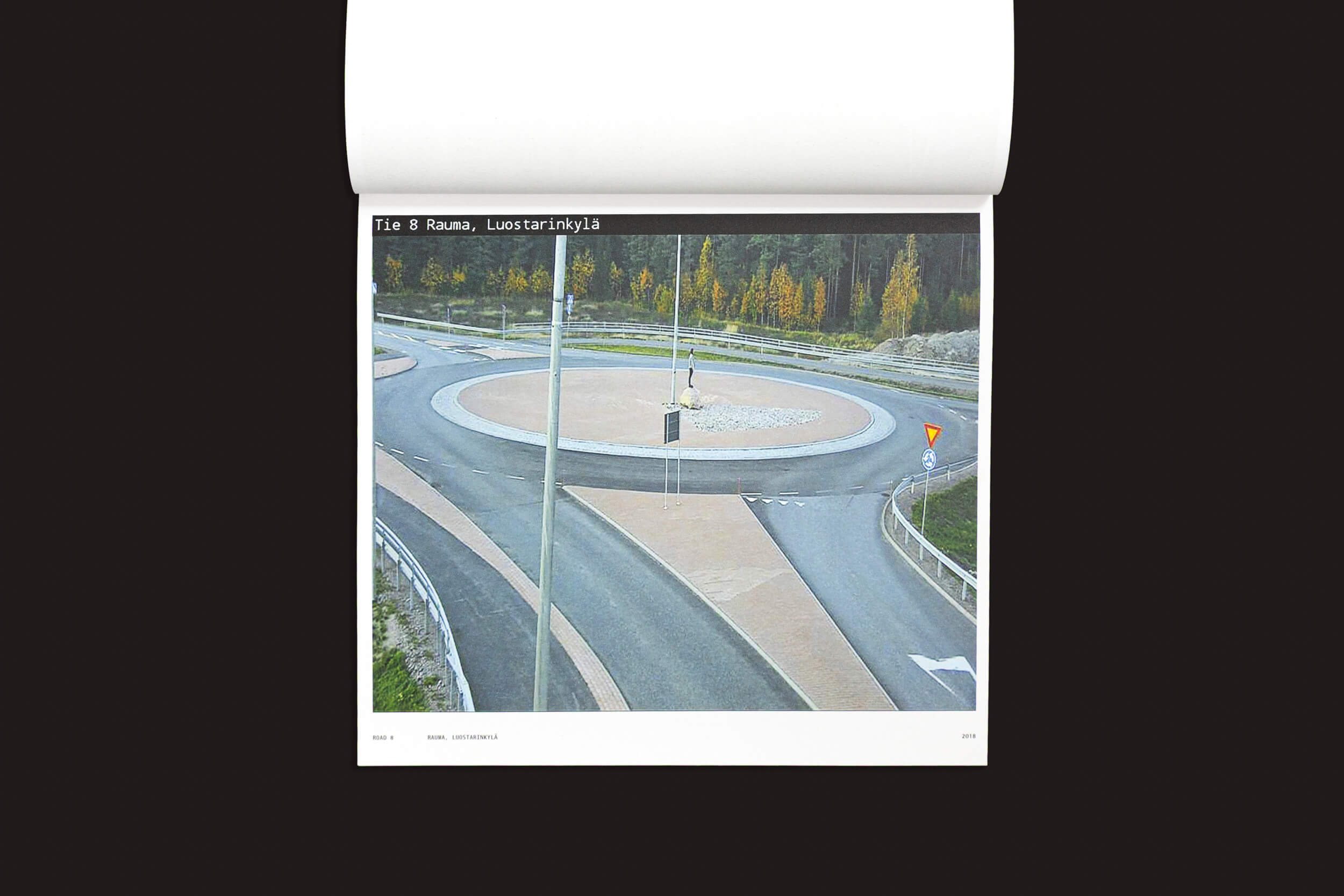 A fotografia alternativa, parte da coleção de Tatu Gustafsson "I on the Road / Weather Camera Self-Portraits, 2012-2021" da Fw:Books, retrata uma rotatória com estradas ramificadas. Um poste e um poste de luz ficam em meio a uma ilha emoldurada por árvores em tons de outono, perto de uma placa com os dizeres "Tie 8 Rauma, Luostarinkylä.