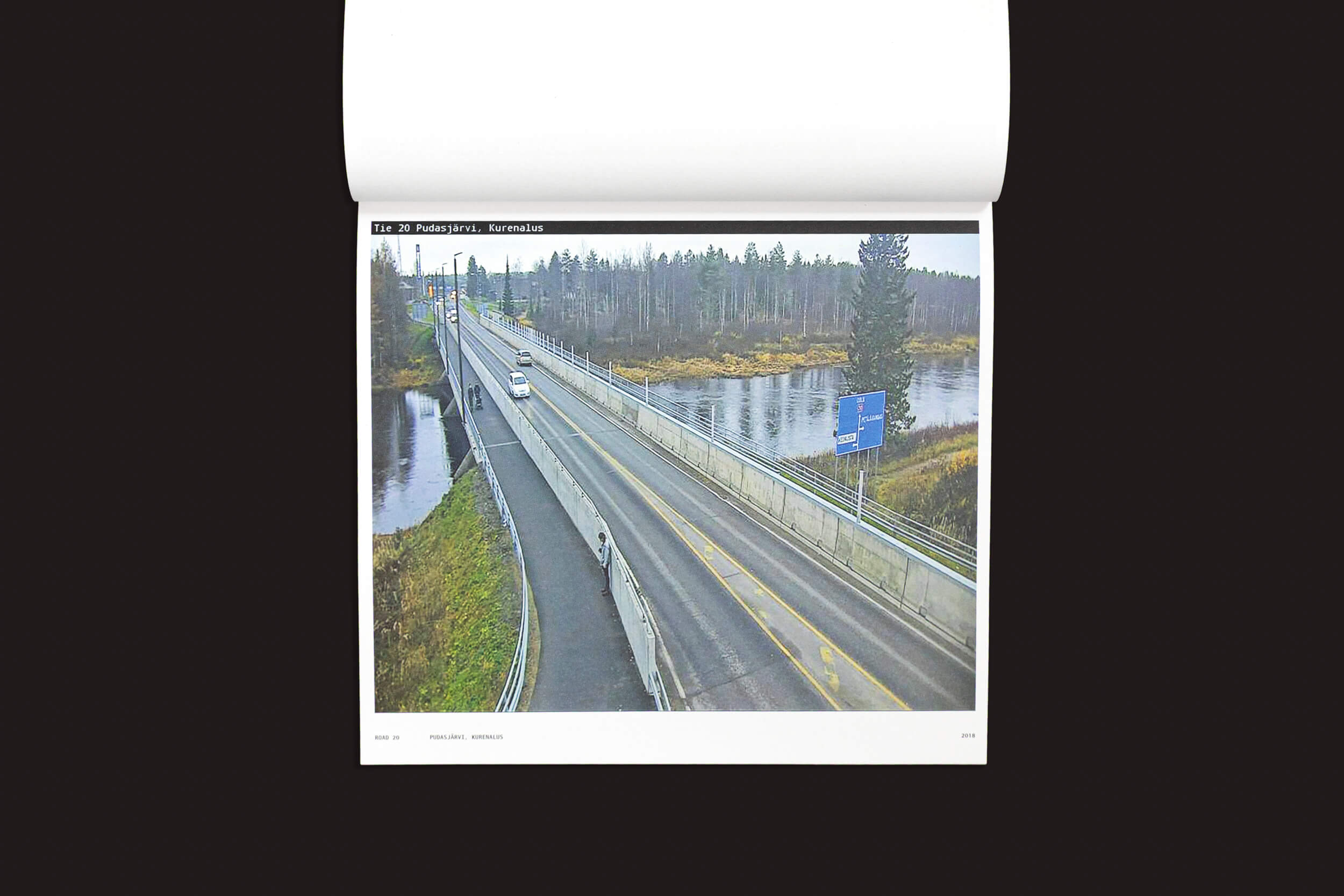 Uma vista aérea de uma ponte rodoviária sobre um rio, cercada por árvores, com um veículo e uma placa azul visíveis. Este cenário é ideal para capturar autorretratos deslumbrantes, como mostrado em "Tatu Gustafsson: I on the Road / Weather Camera Self-Portraits, 2012-2021" por Fw:Books.