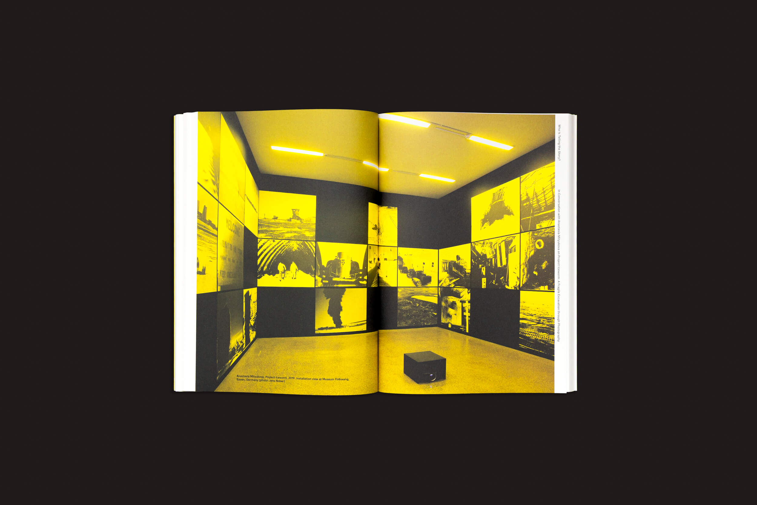 Um livro aberto, "Why Exhibit? vol. 2 - On Curating Photography" da Fw:Books, revela uma sala onde práticas curatoriais ganham vida, com imagens pretas e amarelas nas paredes. Um pequeno banco preto fica no chão enquanto uma iluminação amarela quente realça esta representação fotográfica única de arte curada.