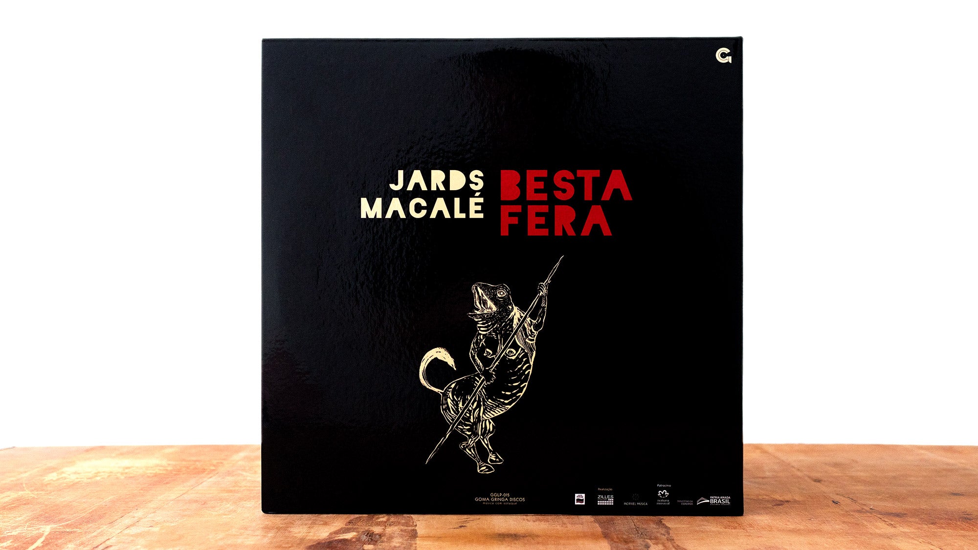Capa preta do álbum Besta Fera de Jards Macalé, com texto dourado, texto vermelho e uma ilustração de um lagarto ou animal em pé empunhando uma lança, sobre uma superfície de madeira com fundo branco.