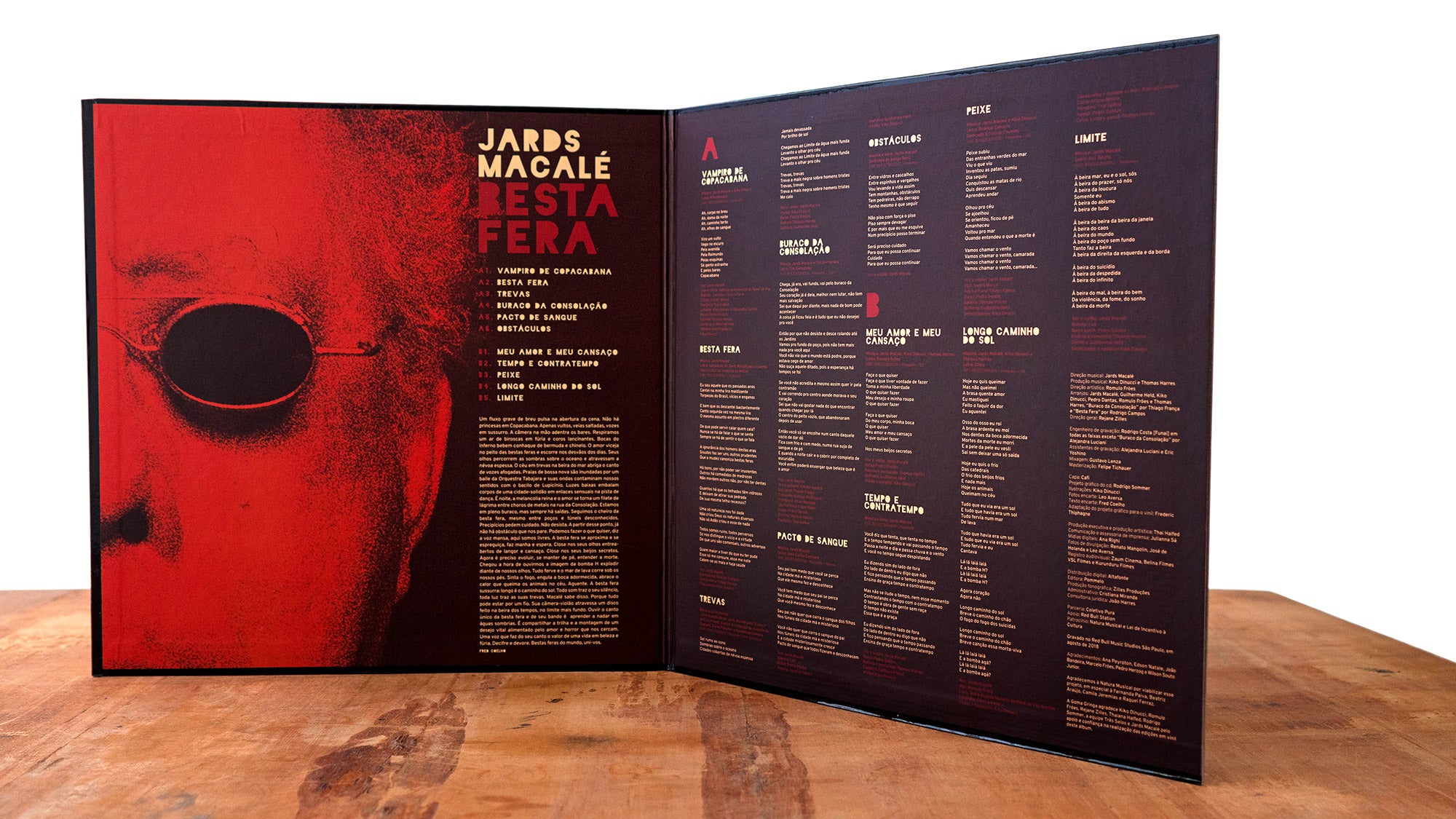 Disco de vinil aberto com capa dupla de Besta Fera, de Jards Macalé, sobre uma superfície de madeira. O lado esquerdo mostra um rosto avermelhado com óculos escuros e informações sobre o álbum; o lado direito lista os títulos das músicas e os créditos em texto dourado e branco sobre um fundo escuro.