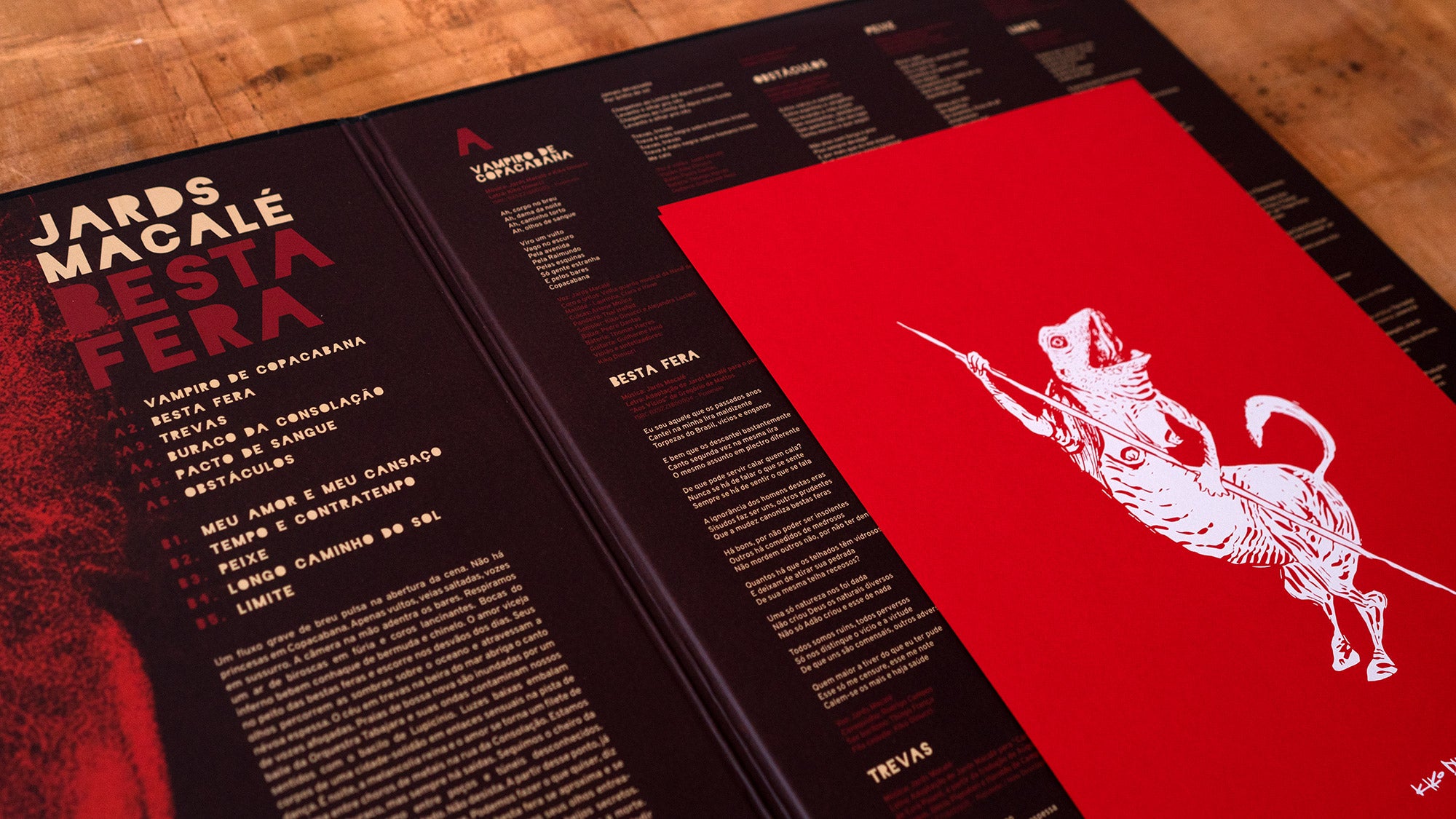 A capa do álbum de vinil Jards Macalés Besta Fera apresenta um texto moderno e em negrito e um encarte vermelho com uma ilustração de uma figura com cabeça de cachorro segurando uma lança, tudo exposto em uma superfície de madeira.