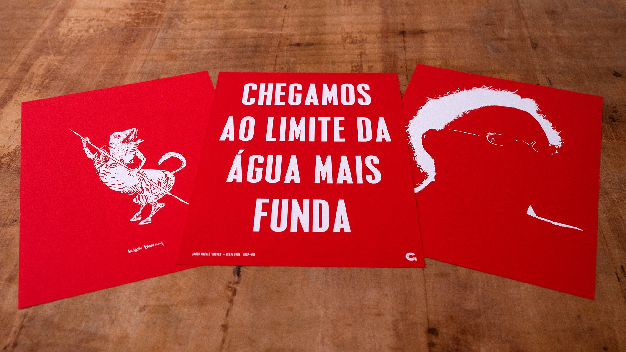 Três cartazes vermelhos sobre uma superfície de madeira: o da esquerda mostra uma ilustração branca de uma pessoa segurando uma lança, o do centro diz CHEGAMOS AO LIMITE DA ÁGUA MAIS FUNDA em português, e o da direita exibe uma silhueta branca de um rosto.
