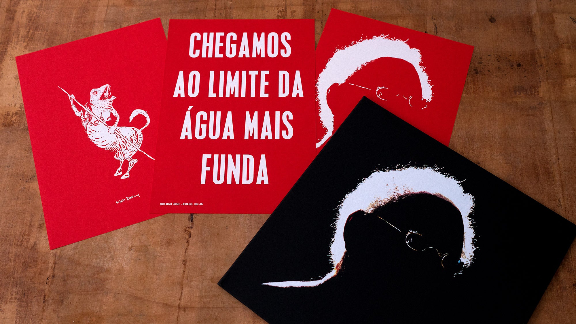 Três peças de arte sobre uma superfície de madeira: uma mostra um cachorro com corpo humano segurando uma lança, outra tem texto em português e as duas últimas retratam a silhueta branca de uma pessoa em fundos vermelho e preto.