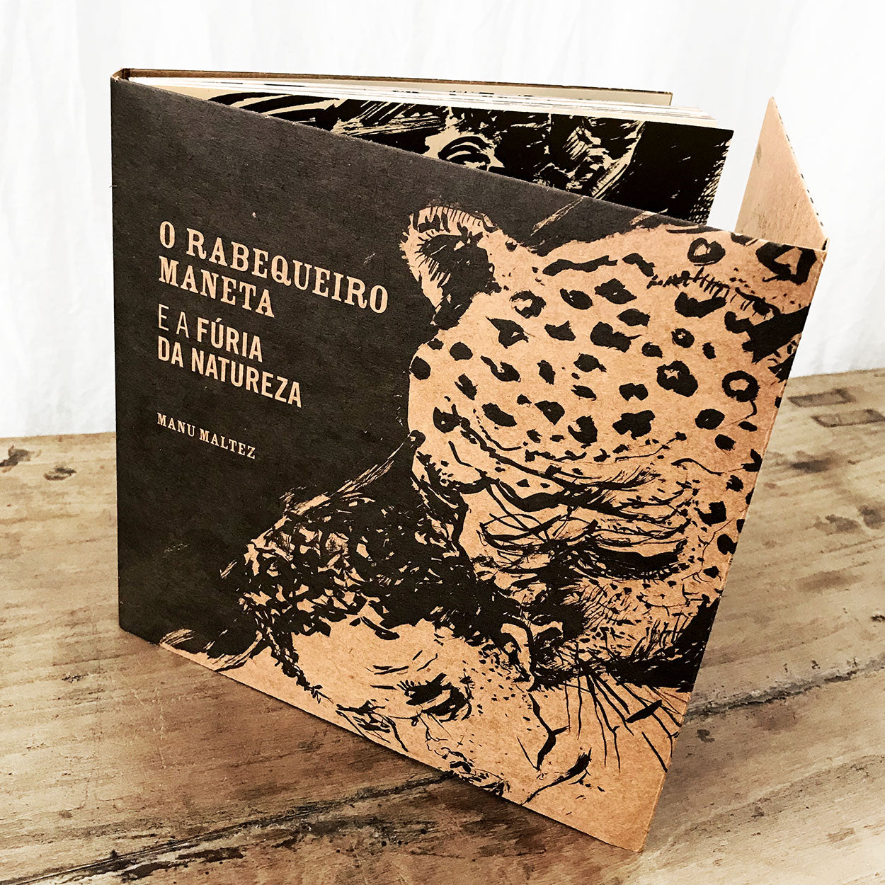 Um livro intitulado O Rabequeiro Maneta e a Fúria da Natureza, de Manu Maltez, que apresenta uma ilustração em preto e marrom de duas onças na capa, em pé sobre uma superfície de madeira.