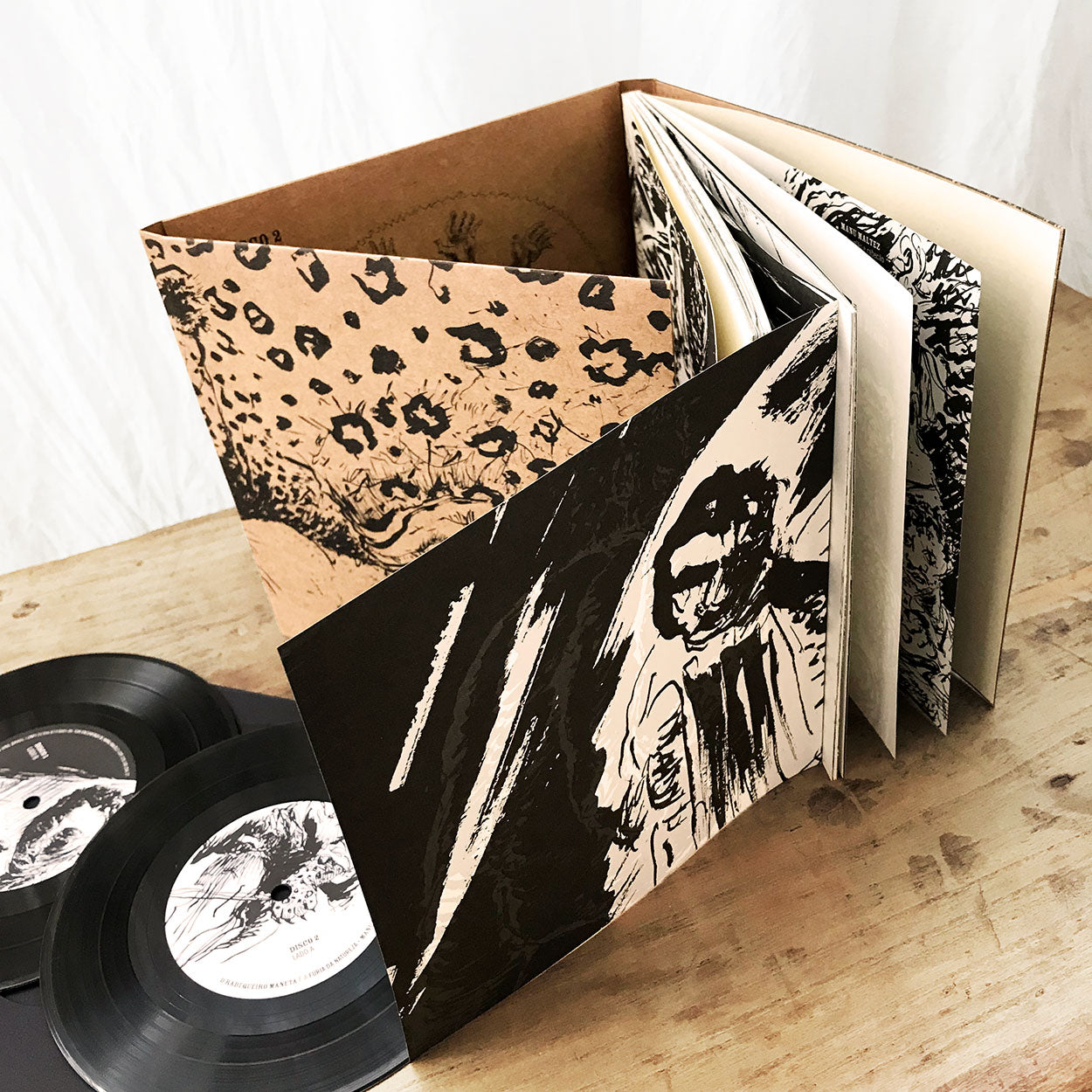 Abra a capa do álbum ilustrada com arte abstrata em preto e bege, em pé sobre uma superfície de madeira, ao lado de dois discos de vinil pretos com etiquetas brancas no centro.