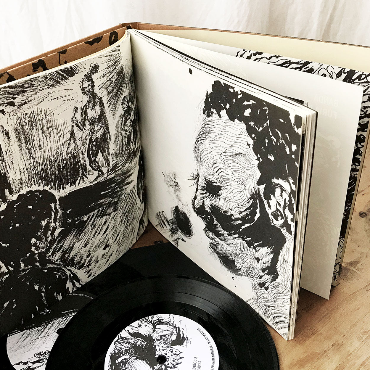 Um livro ilustrado aberto com ilustrações em preto e branco, em estilo esboço, está ao lado de um disco de vinil e sua capa, sobre uma superfície de madeira. As páginas apresentam figuras e rostos humanos expressivos e abstratos.