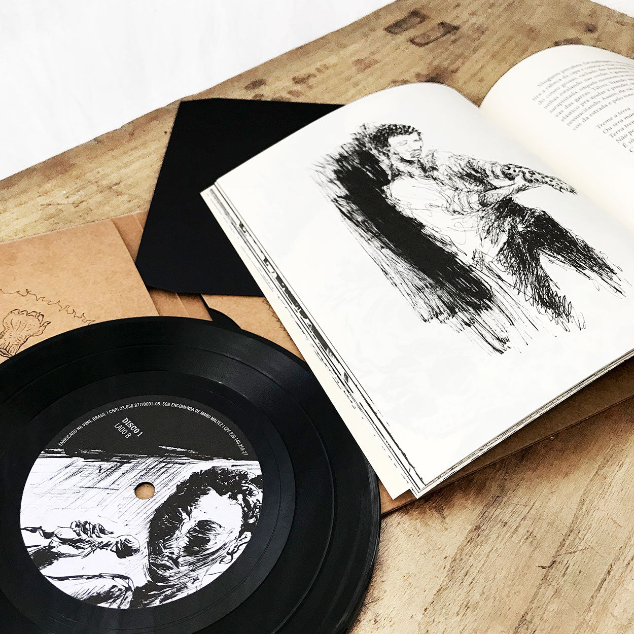 Um disco de vinil com o desenho de um cantor no rótulo repousa sobre uma superfície de madeira ao lado de um livreto aberto com um desenho em preto e branco de um artista segurando um microfone. Envelopes marrons são parcialmente visíveis embaixo.