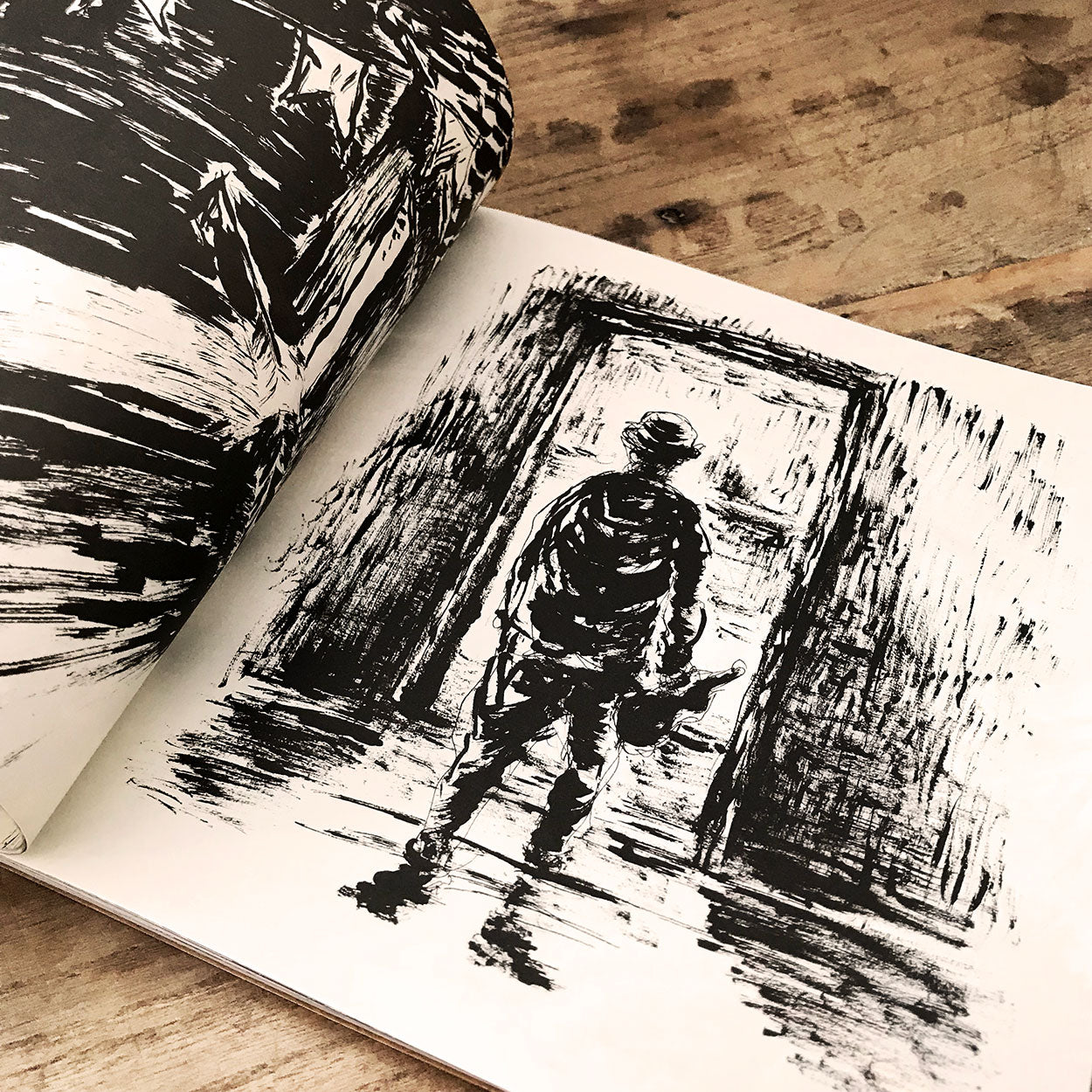 Um esboço em preto e branco mostra uma pessoa em pé diante de uma porta, segurando algo na mão, com sombras e linhas texturizadas criando uma atmosfera dramática e melancólica. O livro está aberto sobre uma superfície de madeira.