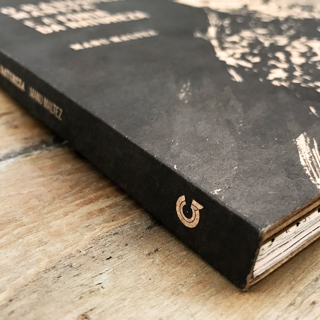 Close-up do canto de um livro preto com texto branco na capa, sobre uma superfície de madeira. A lombada apresenta um pequeno logotipo circular próximo à parte inferior. As páginas do livro estão ligeiramente irregulares.