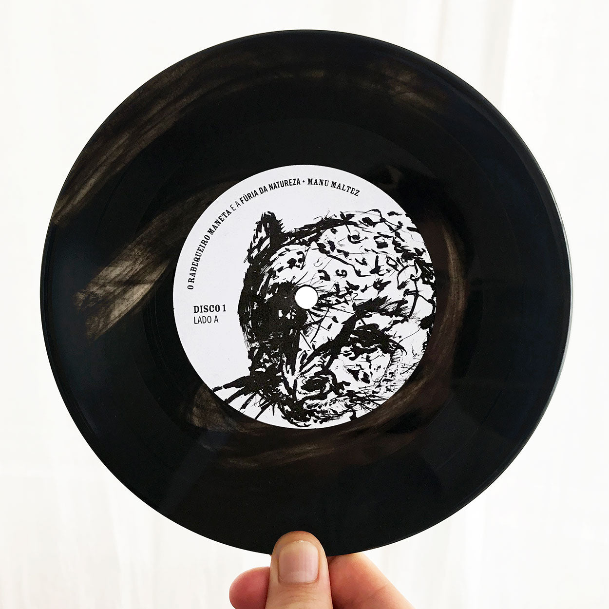 Uma mão segura um disco de vinil preto com uma etiqueta branca com um desenho abstrato em tinta preta do rosto de um animal. O rótulo diz: O jardim dos habitantes do fundo da natureza • Manu Maltez e Disco 1 Lado A.