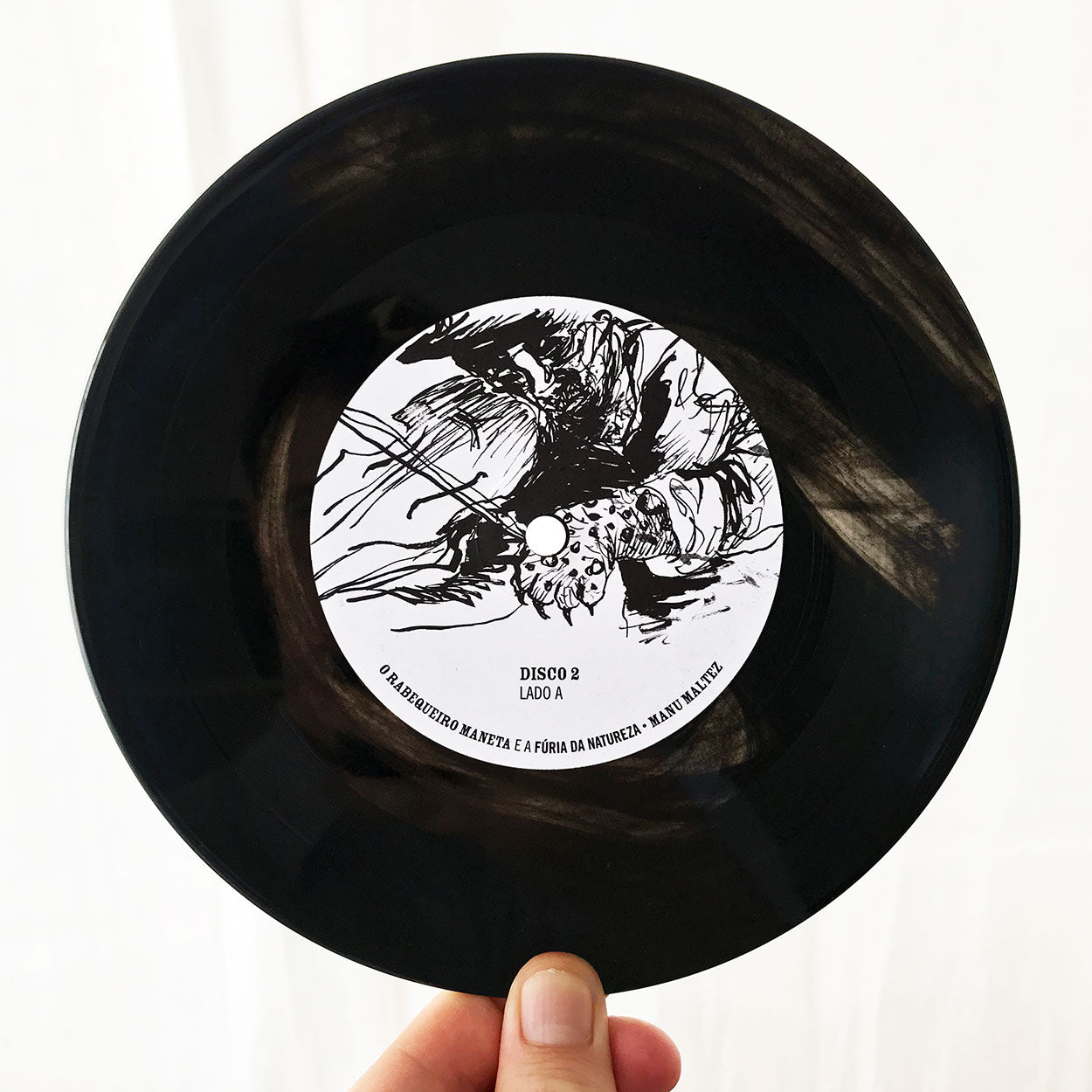 Uma mão segura um disco de vinil preto com uma etiqueta branca com arte abstrata em preto e texto em português, incluindo “DISCO 2 LADO A” e um texto menor adicional ao redor da borda da etiqueta.