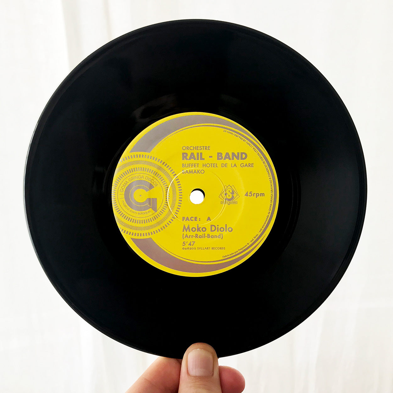 Uma mão segura um disco de vinil preto com uma etiqueta amarela que diz "Orchestre Rail Band, Buffet Hotel de la Gare, Bamako". O disco se chama "Moko Diolo" e toca a 45 RPM. O fundo é liso e branco.