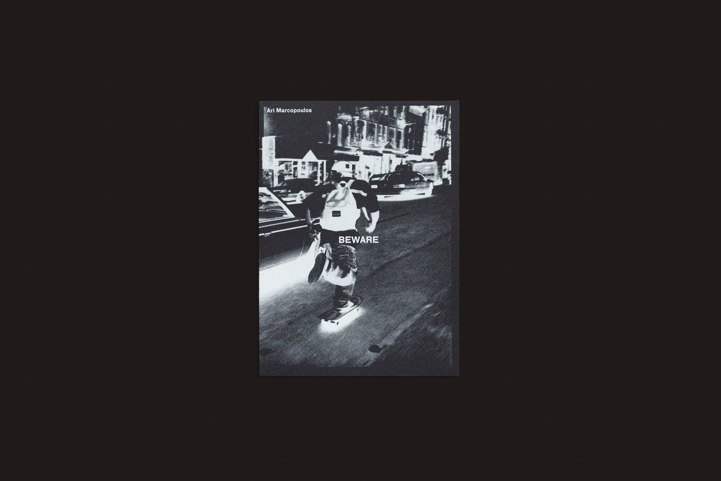 Uma foto em preto e branco captura um skatista em uma movimentada rua da cidade à noite, lembrando o estilo de Ari Marcopoulos. A palavra BEWARE paira sobre eles, com prédios e carros se misturando ao fundo, evocando a essência dos skatistas em Nova York. Descubra mais em Ari Marcopoulos: Beware da Roma Publications.