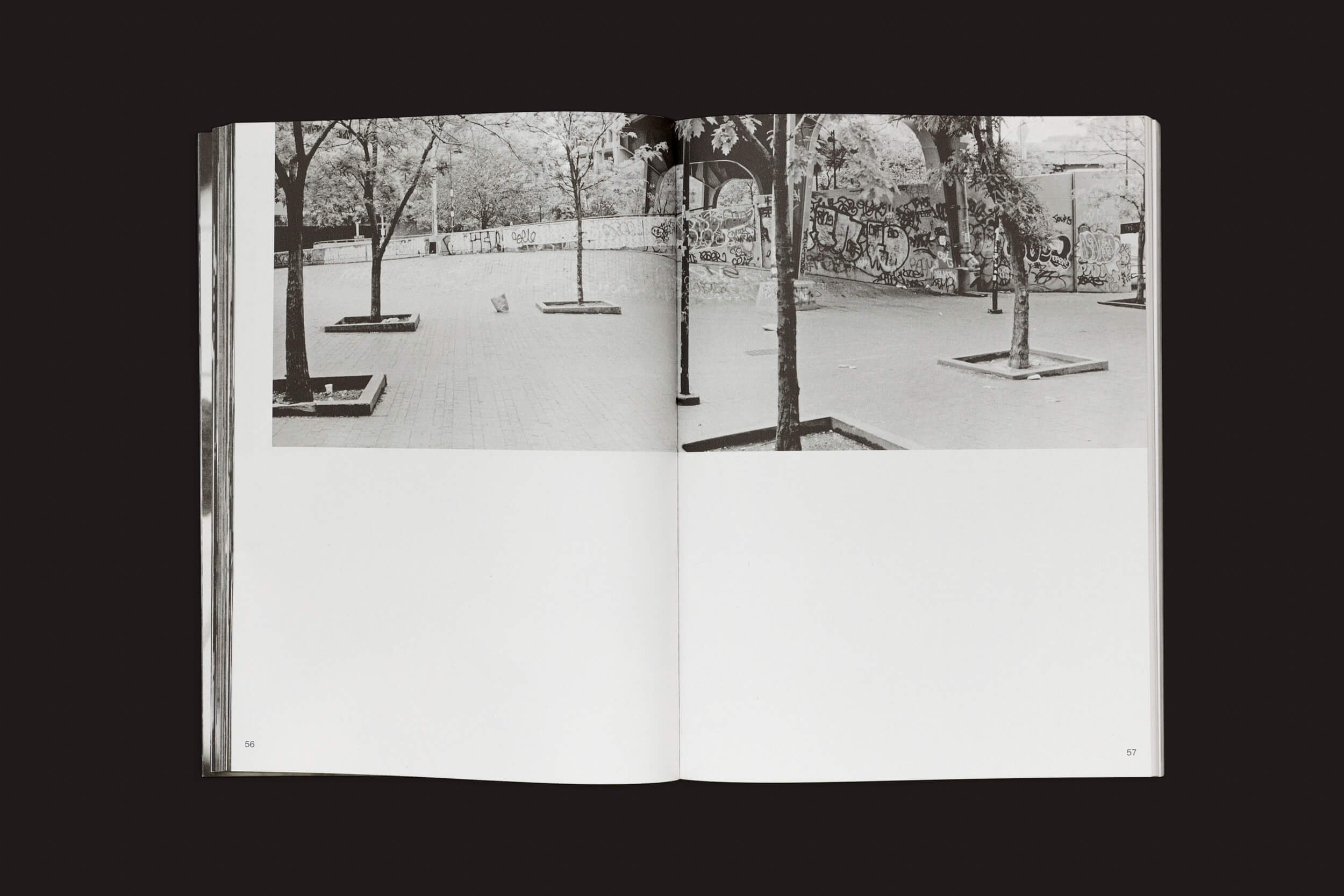 Em Ari Marcopoulos: Beware da Roma Publications, uma página aberta exibe a foto em preto e branco de Marcopoulos de uma rua arborizada e adornada com grafites, ecoando skatistas em Nova York. A continuação da imagem no canto superior direito mostra mais árvores e muros, com o resto quase todo em branco.