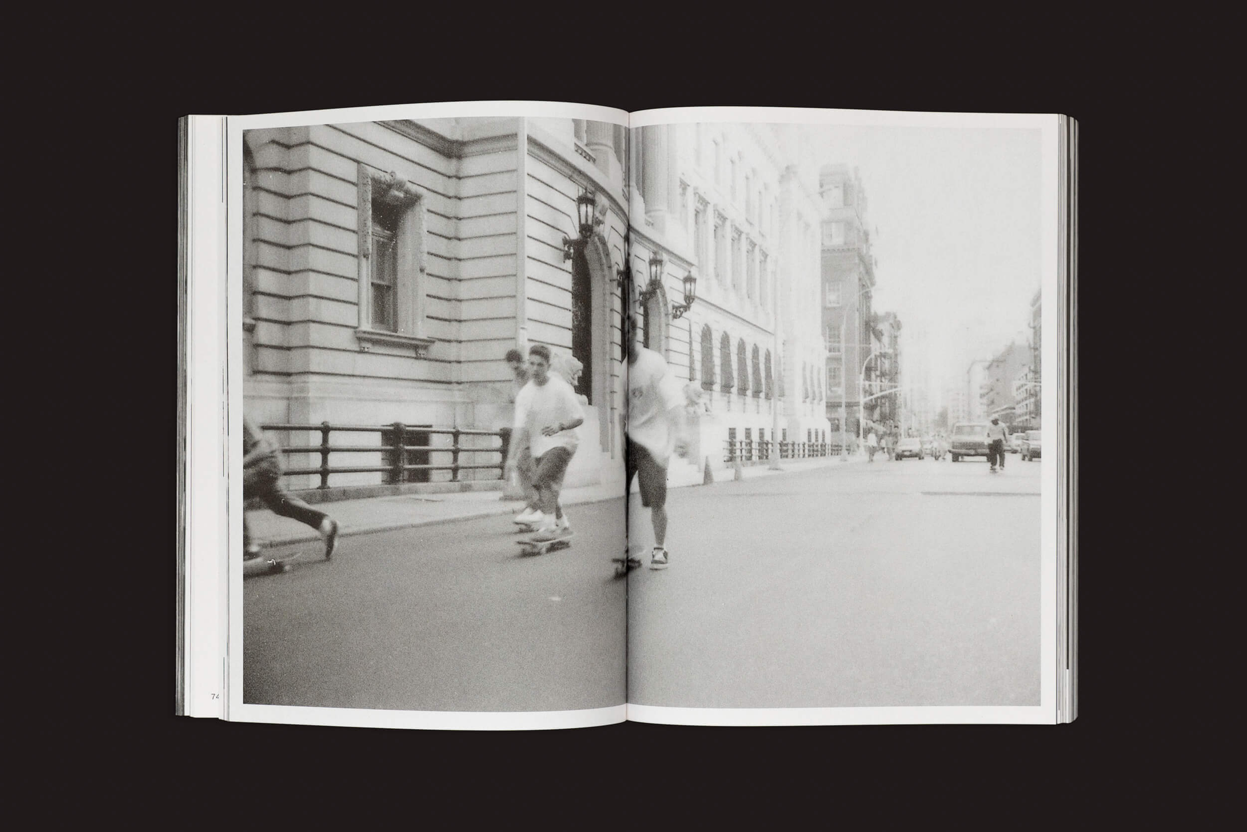 Uma foto do livro Ari Marcopoulos: Beware, da Roma Publications, mostra skatistas em Nova York andando de skate por uma rua com prédios antigos, capturando o movimento da vida urbana com figuras borradas e uma estrada vazia ao fundo.