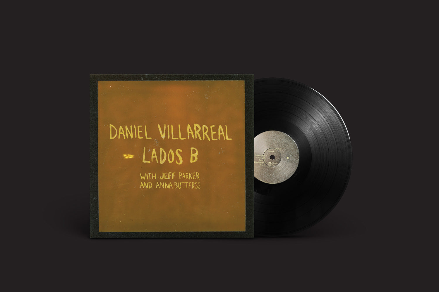 O disco de vinil, "Daniel Villarreal: Lados B" da International Anthem Recording Company, apresenta uma capa com texto manuscrito e lista os colaboradores Jeff Parker e Anna Butterss. A arte vibrante dá vida ao fundo marrom-dourado quente enquanto o disco aparece ao lado dele.