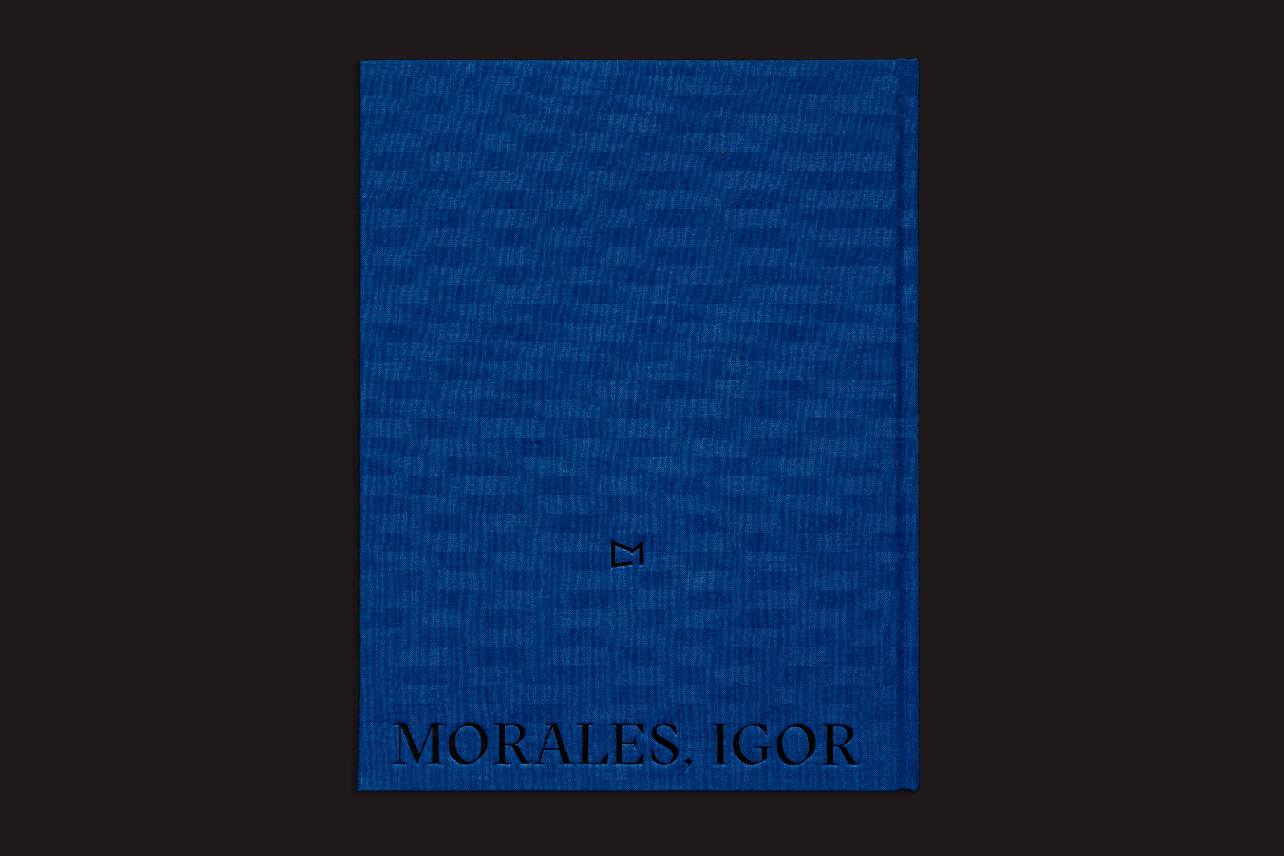 Um livro de capa dura azul intitulado "Igor Morales: Em um Tritão me transformarei eventualmente" da Editora Madalena, com logotipo central minimalista e MORALES, IGOR em texto preto - um design evocativo que sugere emoções através da fotografia.