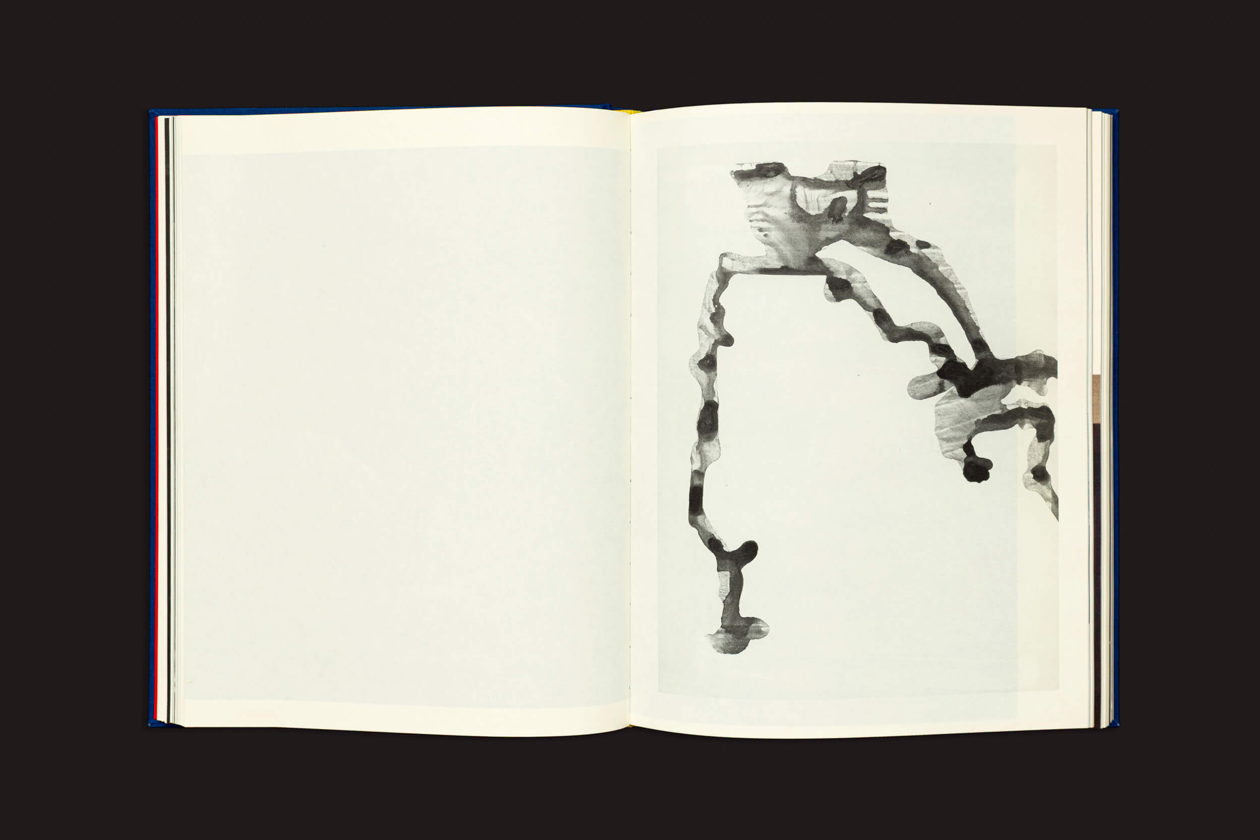 O livro "Igor Morales: Em um Tritão me transformarei eventualmente", da Editora Madalena, mostra uma página em branco à esquerda e uma página à direita com arte abstrata em tinta preta e cinza, evocando emoções e incertezas sobre um fundo preto rígido.