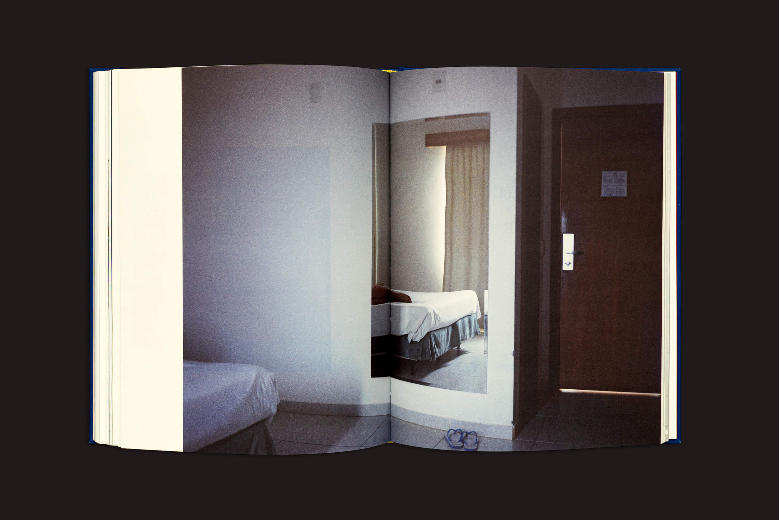 O livro aberto, "Igor Morales: Em um Tritão me transformarei eventualmente", da Editora Madalena, revela a fotografia de um modesto quarto de hotel – cama, paredes brancas, porta marrom e espelho – capturando emoções em detalhes do cotidiano, como sandálias no piso de cerâmica.