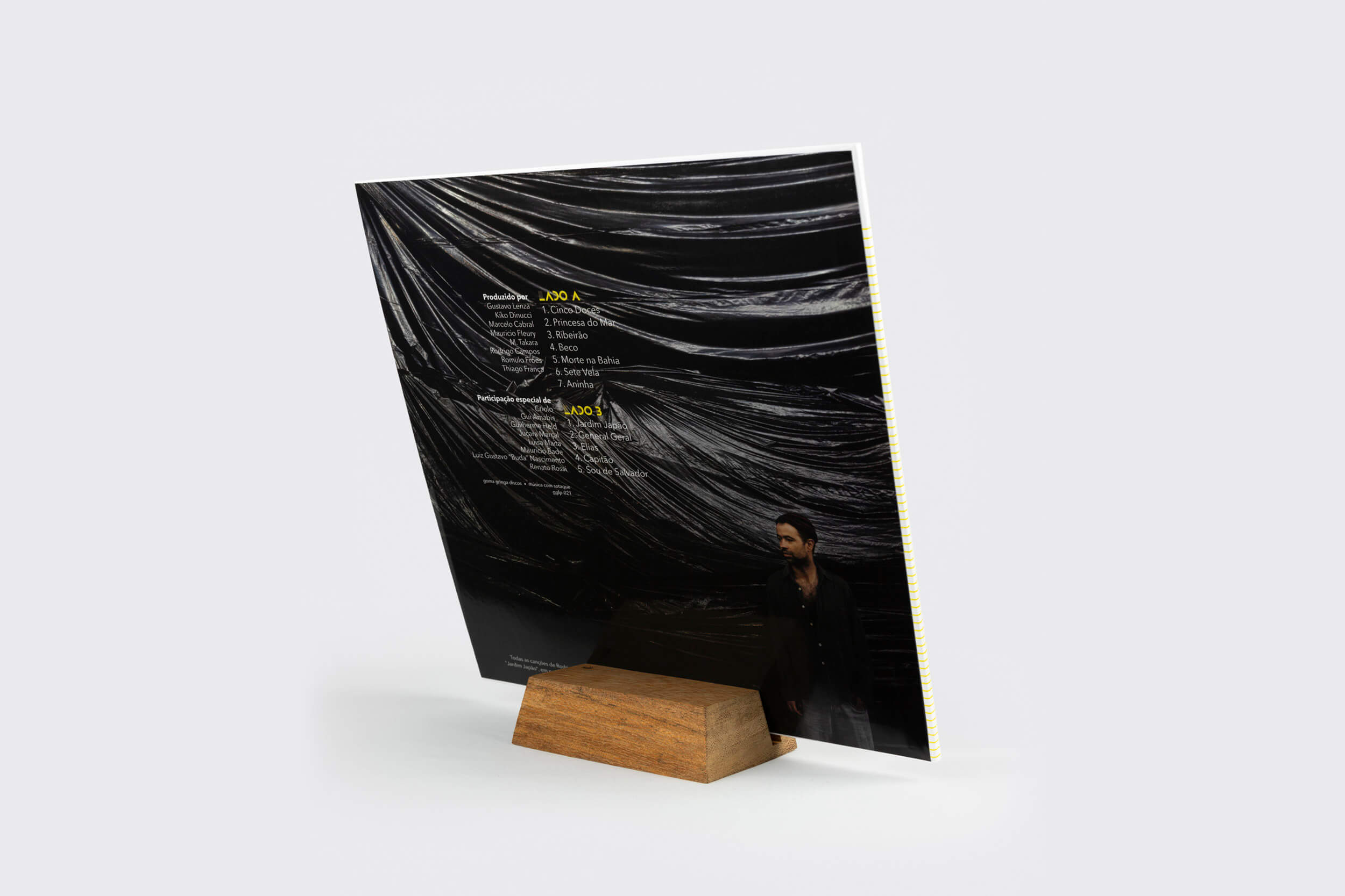 O Display para LP da Goma Gringa apresenta uma capa de disco de vinil em posição vertical dentro do display de madeira. A capa da edição limitada apresenta um design de tecido brilhante e drapeado e uma lista de faixas amarela na parte de trás, com uma pessoa em roupas escuras parcialmente visível na parte inferior.