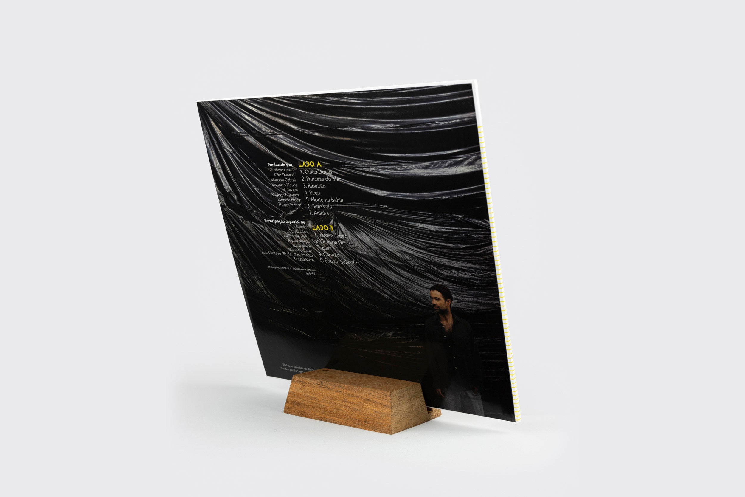 O LP Goma Gringa Display apresenta uma capa de edição limitada em um display de madeira. A contracapa lista os títulos das músicas e os créditos sobre um tecido escuro, com uma pessoa de preto parcialmente visível no canto inferior direito.
