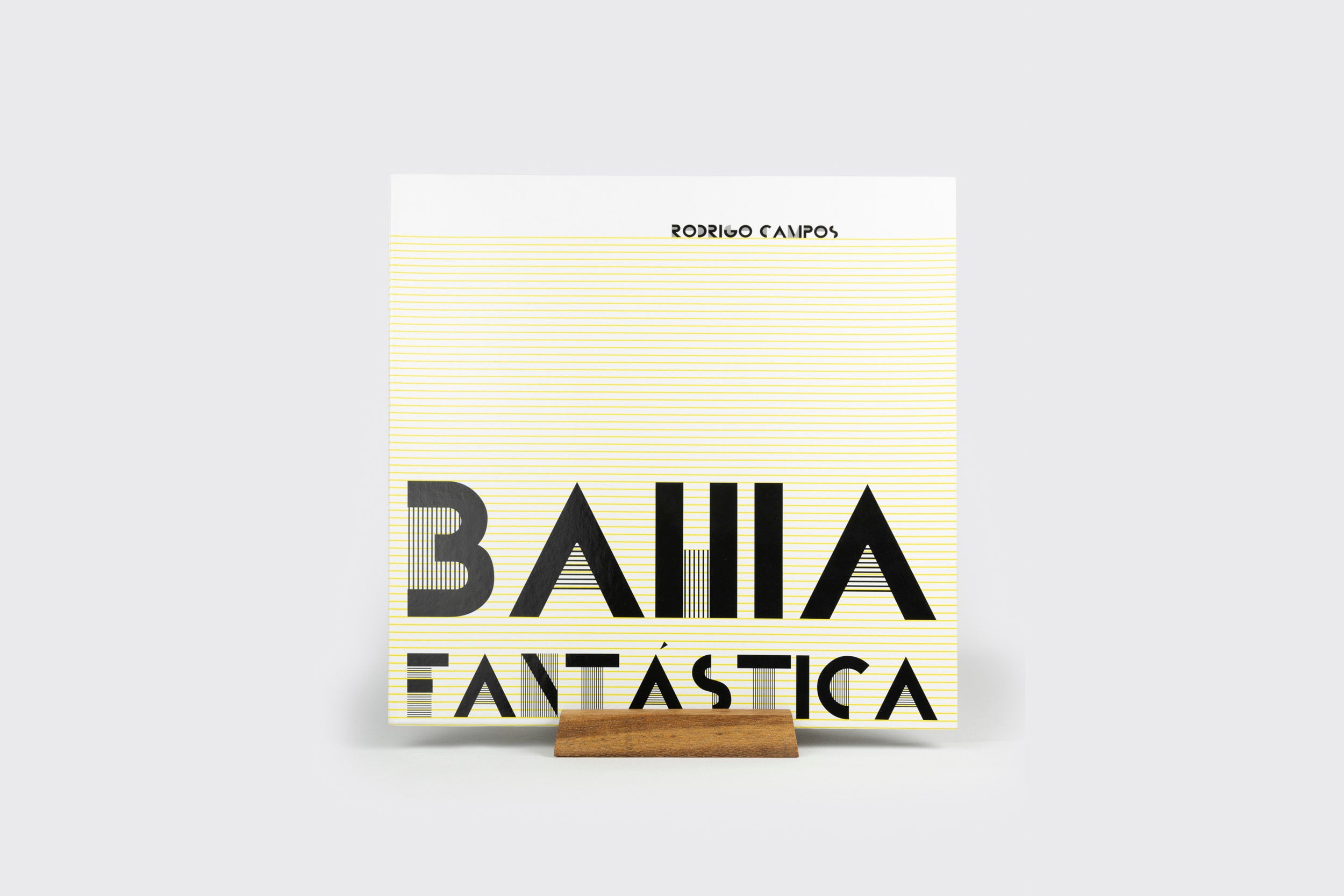 Uma capa quadrada de álbum LP intitulada Bahia Fantástica, de Rodrigo Campos, com letras geométricas pretas em negrito sobre linhas amarelas, é exibida em um suporte de exposição de LPs Goma Gringa feito de madeira simples.