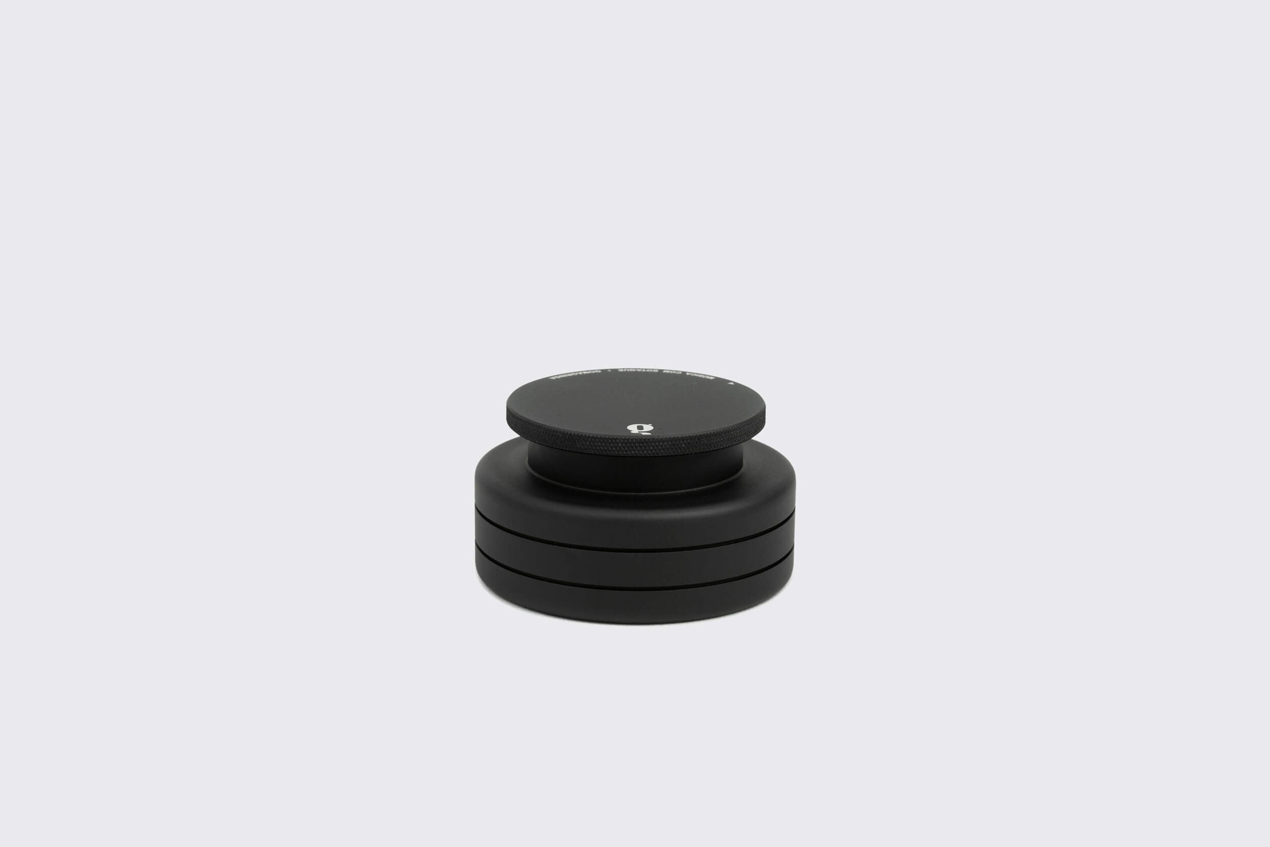 A edição limitada Estabilizador de Disco: Modelo A da Goma Gringa apresenta um design redondo preto elegante com acabamento fosco em alumínio anodizado. Seu formato cilíndrico com duas seções em camadas se destaca elegantemente em um fundo branco simples, destacando sua aparência limpa e moderna.