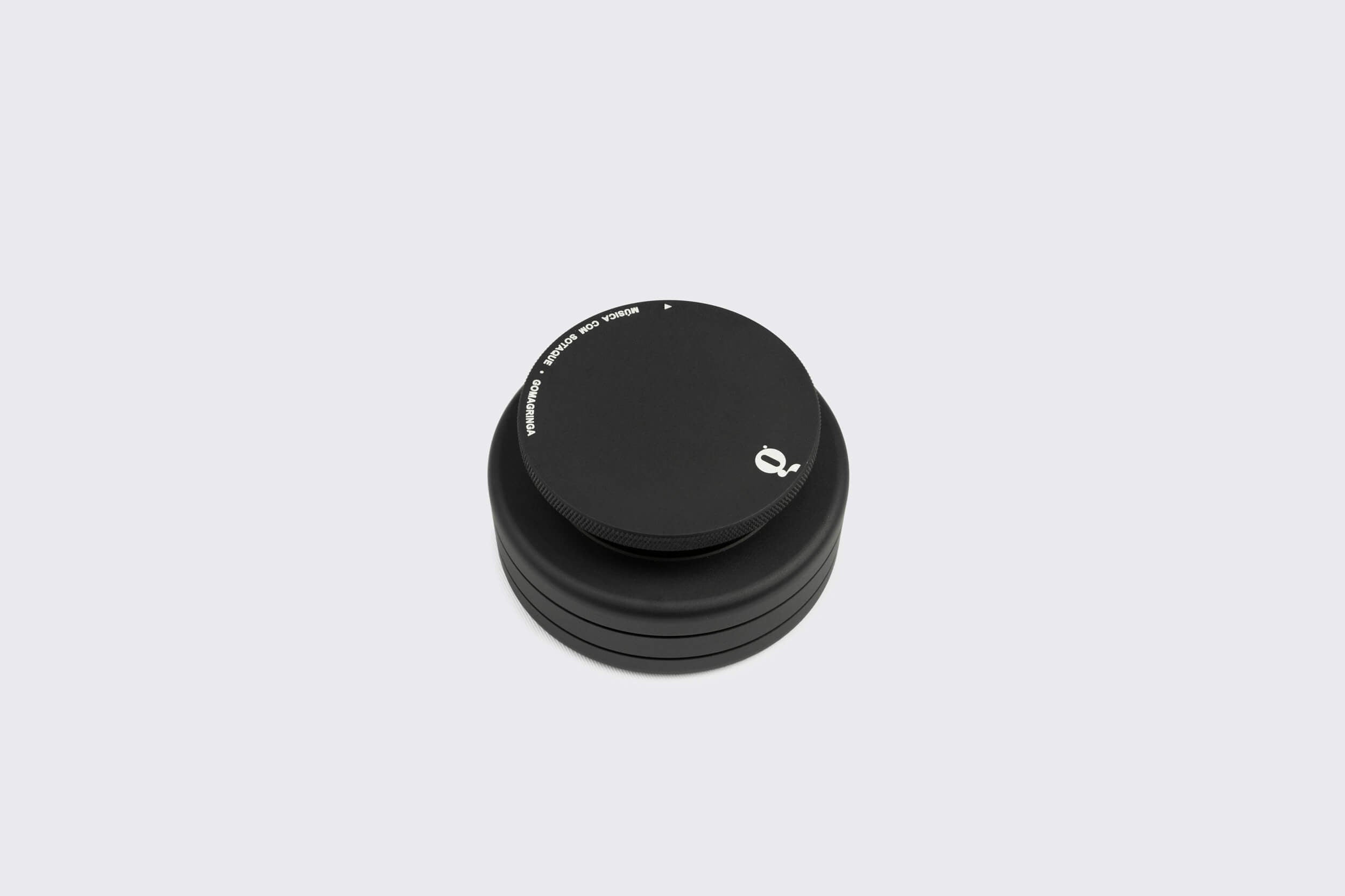 Uma pilha de Estabilizador de Disco: Modelo A preto da Goma Gringa, com um logotipo circular branco, lembrando um peso de toca-discos elegante, é elegantemente colocada sobre um fundo cinza claro simples.