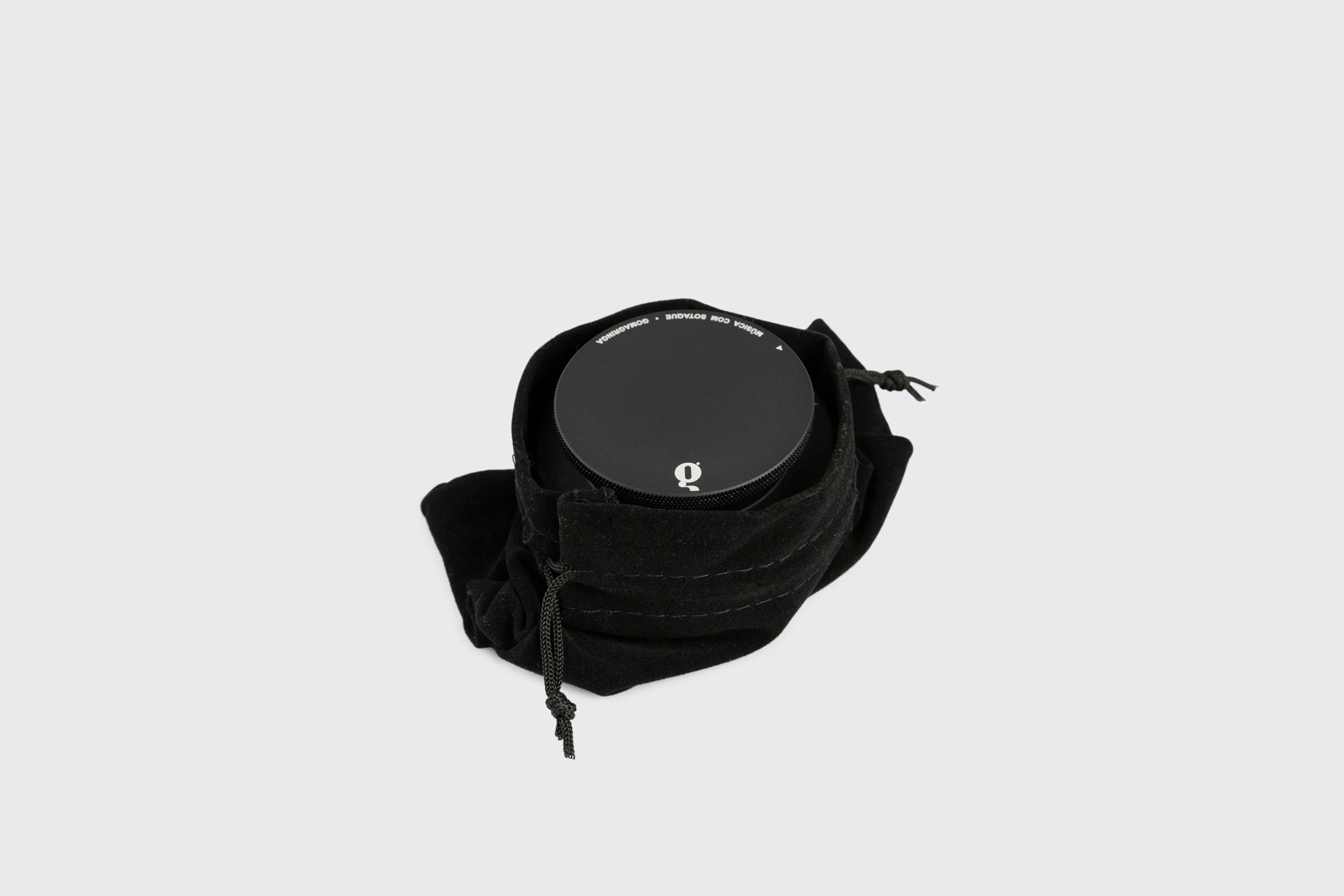 Um pote redondo preto com tampa, semelhante à Goma Gringa Estabilizador de Disco: Modelo A, parcialmente envolto em uma bolsa macia com cordão preto, fica sobre um fundo cinza claro simples.