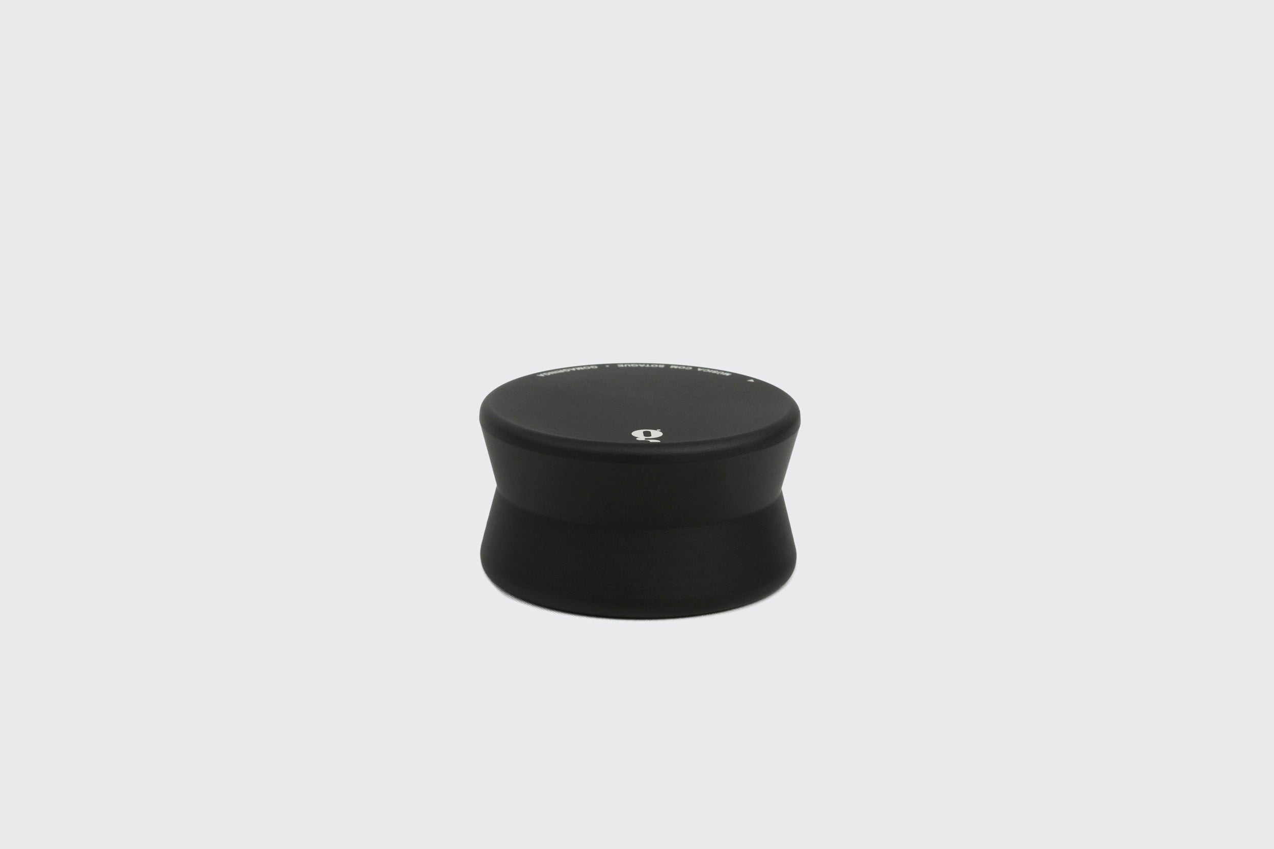 Um Estabilizador de Disco: Modelo B, preto e cilíndrico, da Goma Gringa, com base mais larga e topo plano, repousa sobre um fundo cinza claro. Seu topo exibe um pequeno logotipo branco no centro, indicando um lançamento de edição limitada.