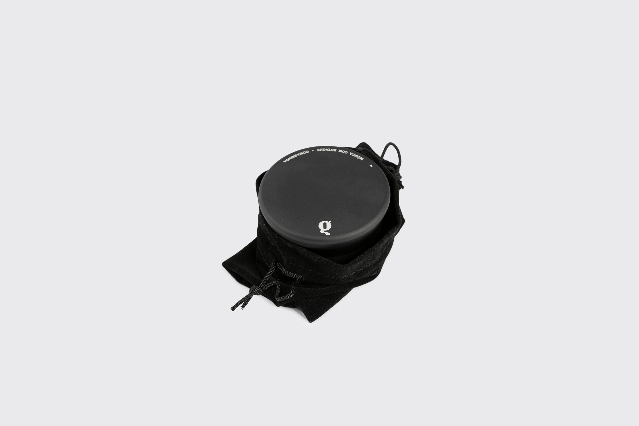 Um recipiente redondo preto com um logotipo branco da Goma Gringa na tampa, parcialmente escondido em uma bolsa preta com cordão, sobre um fundo cinza claro — perfeito para armazenar seu Estabilizador de Disco: Modelo B.