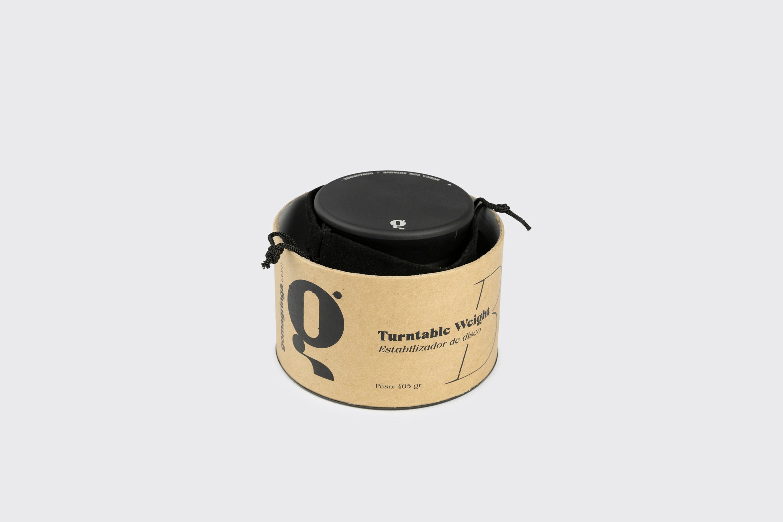 Um Estabilizador de Disco: Modelo B preto da Goma Gringa, feito de alumínio anodizado, repousa parcialmente em uma capa redonda de papelão rotulada "Turnttable Weight" com texto extra e um "Q", sobre um fundo cinza claro.