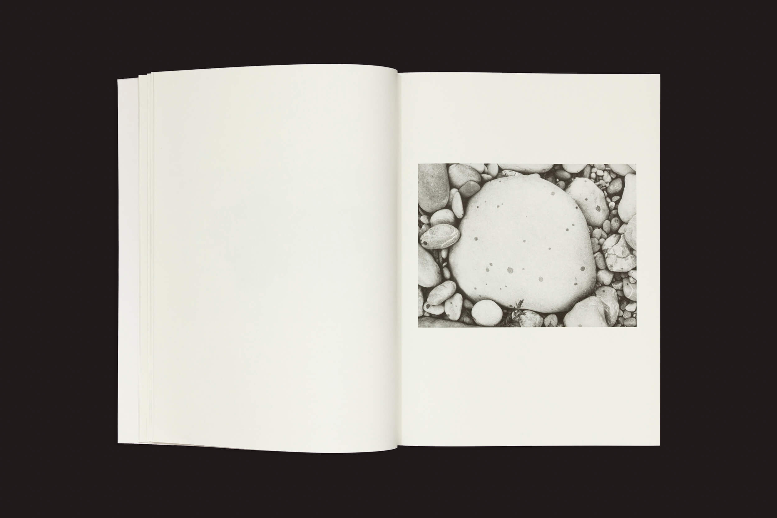 Jochen Lempert: Paare / Pairs, da Roma Publications, está aberto sobre uma superfície escura, exibindo fotos em preto e branco de seixos ao redor de uma grande pedra na página direita, enquanto a página esquerda está em branco.
