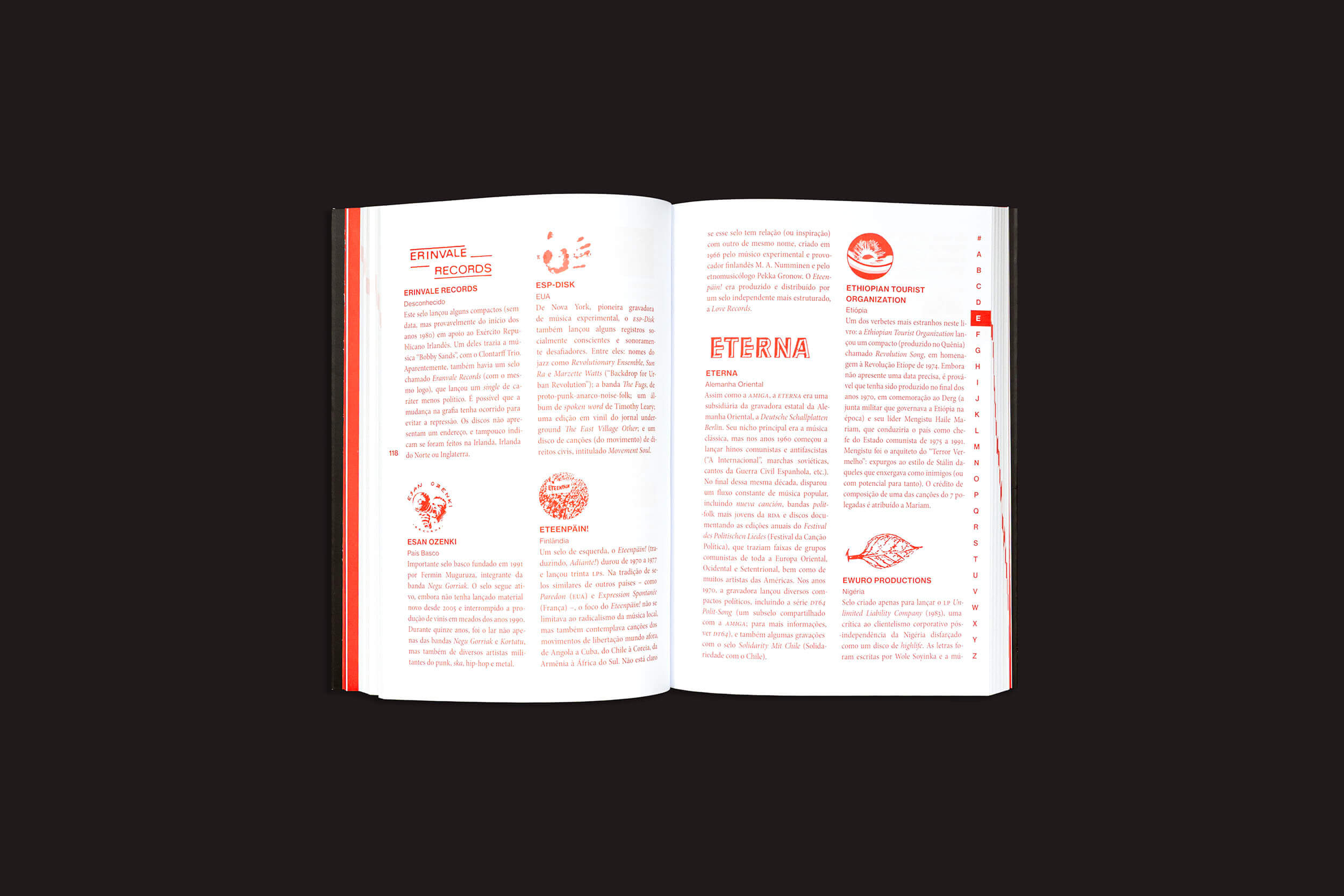 O livro aberto "Josh MacPhee: Radical Records" de sobinfluencia apresenta uma página branca com texto em vermelho e preto, ecoando a energia vibrante da música independente. Pequenas imagens, logotipos e ícones adicionam toques decorativos, enquanto o fundo preto simples reflete a profundidade das discos de vinil.