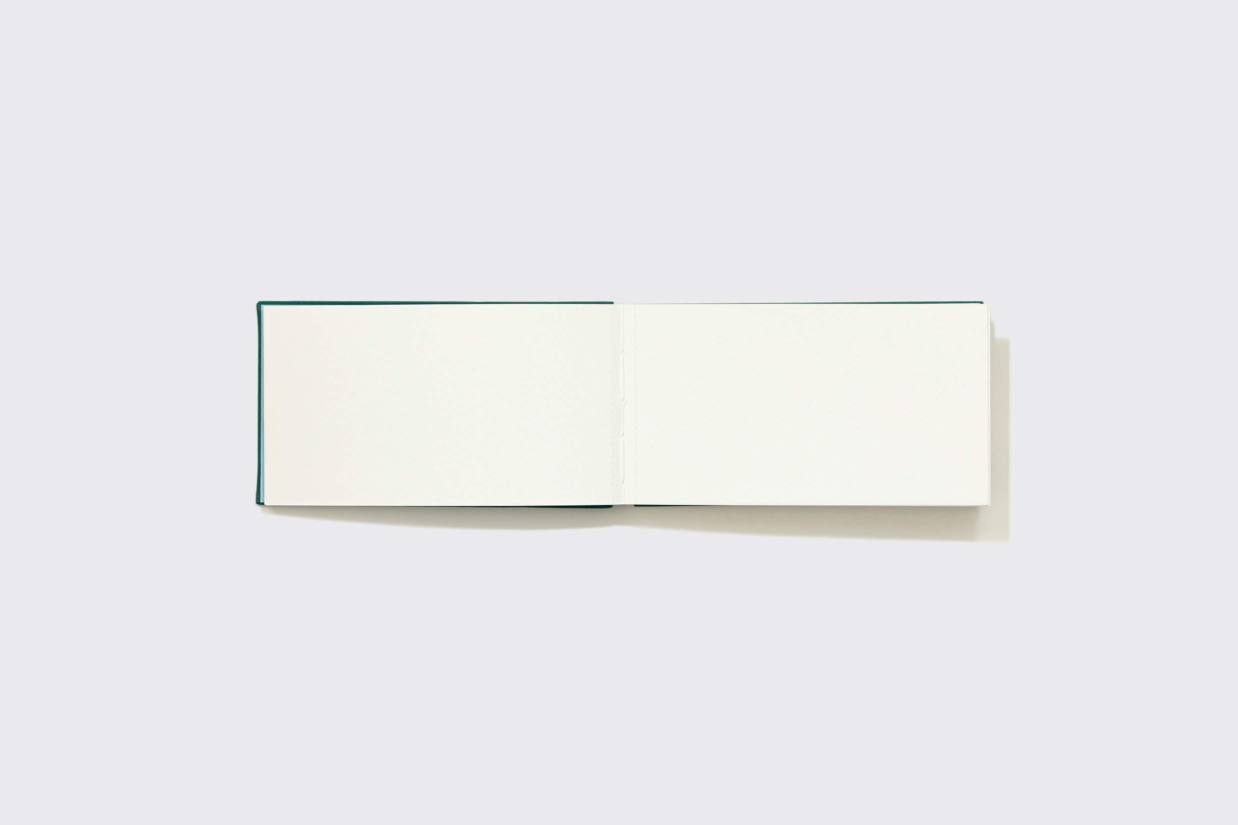 O Caderno Aquarela Pocket: Alecrim da Schizzibooks está aberto sobre uma superfície branca, revelando duas páginas em branco para seus pensamentos. Sua capa verde escura acrescenta sofisticação enquanto a iluminação suave projeta uma sombra, convidando suas ideias a se derramarem na tela imaculada.