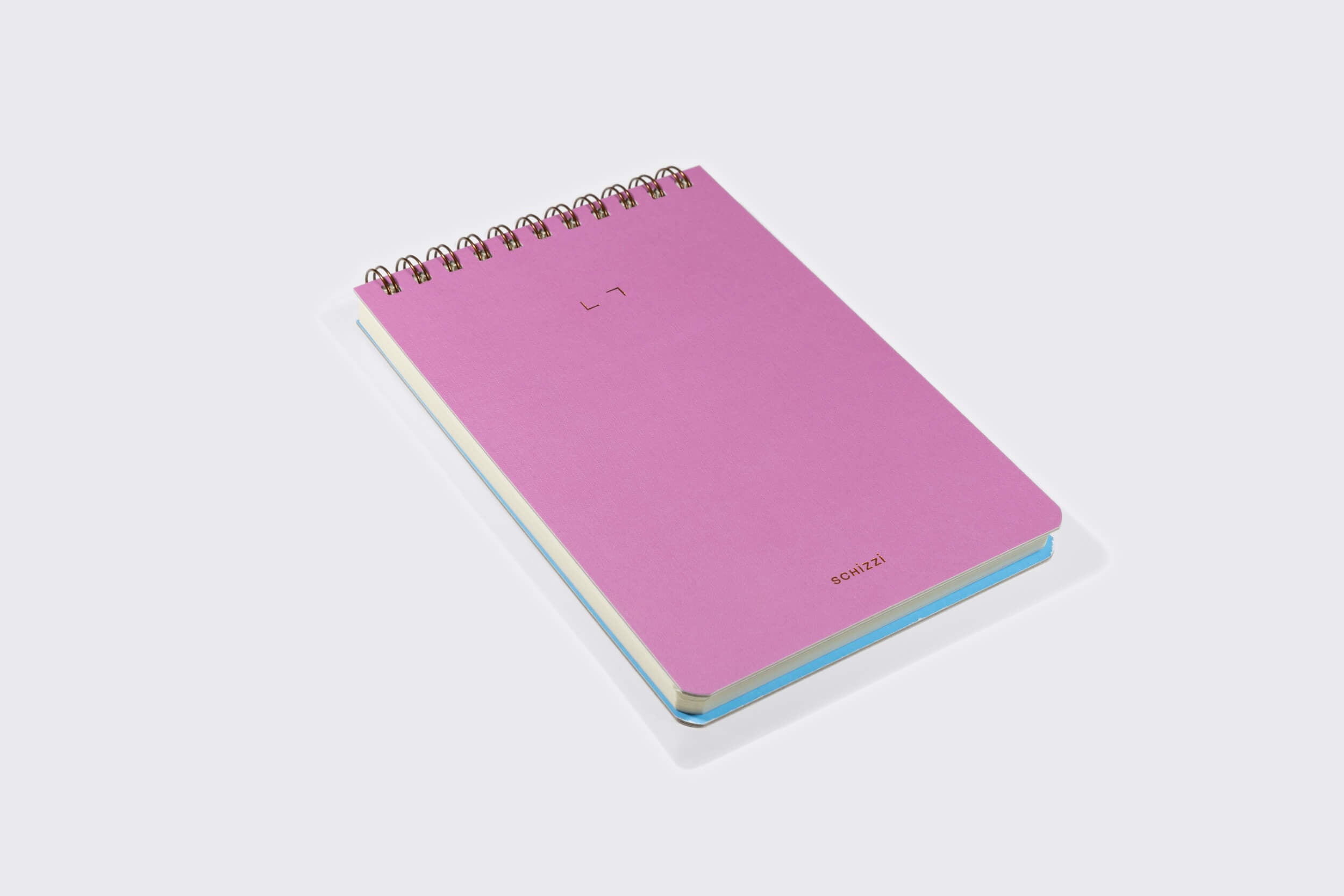 The Caderno Reporter: Mirtilo da Schizzibooks, um caderno espiral rosa feito à mão do Brasil, fica em uma superfície cinza claro. Seu design minimalista inclui um pequeno texto na parte superior e inferior da capa com páginas de papel pólen macio elegantemente espreitando por baixo.