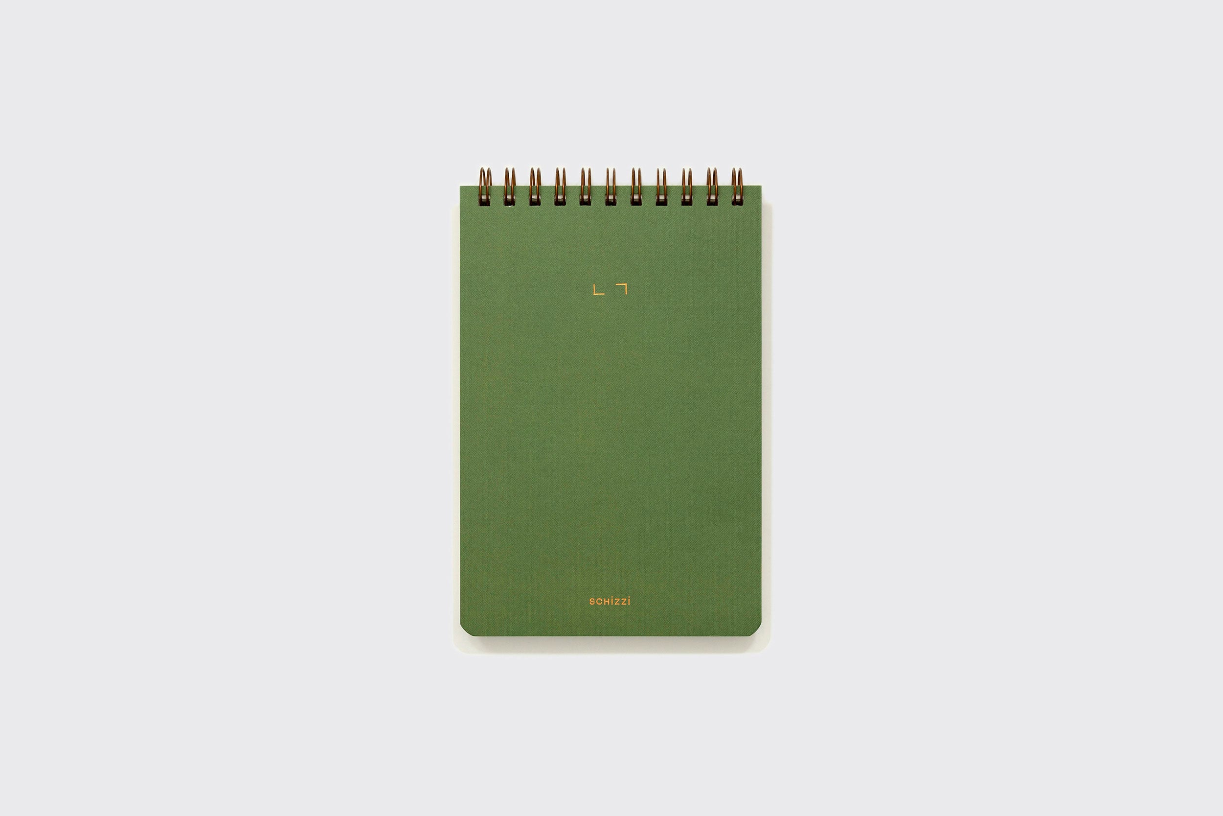 Um Caderno Reporter verde da Schizzibooks, encadernado em espiral com texto dourado na capa e feito à mão no Brasil, fica em pé sobre um fundo cinza claro simples.