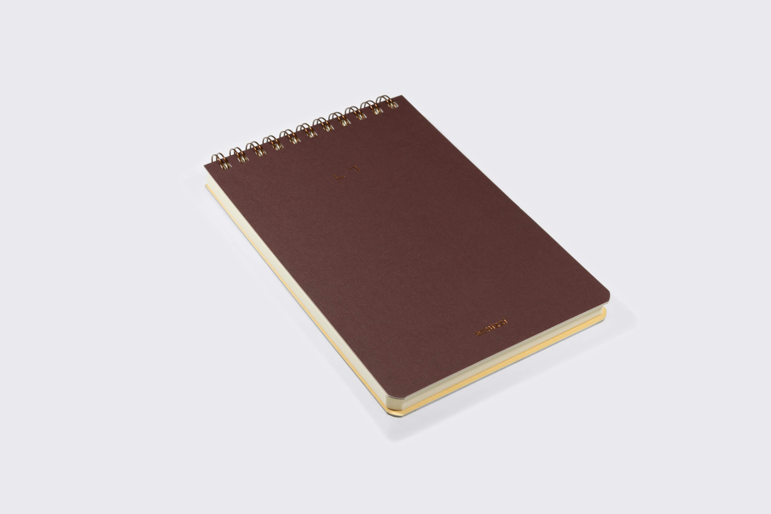 Um Caderno Reporter: Savana encadernado em espiral marrom da Schizzibooks com capa texturizada repousa sobre uma superfície cinza-claro. O caderno fechado exibe suas espirais douradas e páginas sem ácido, meticulosamente produzidas manualmente.