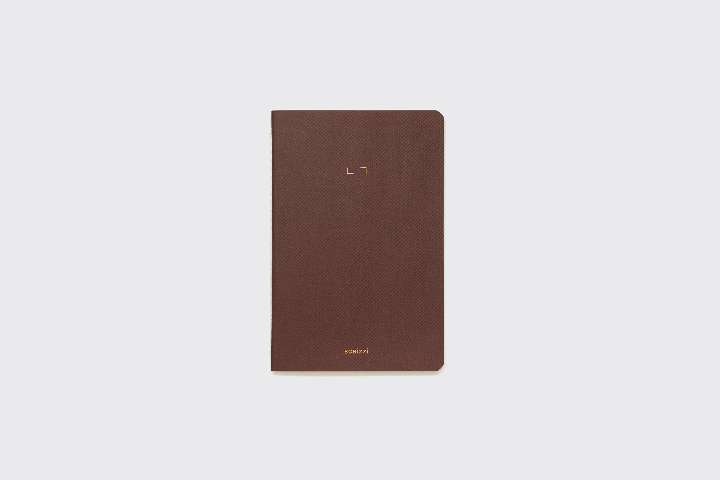 O Caderno Flexível da Schizzibooks é um caderno marrom (15x22cm) feito à mão com cantos arredondados e texto dourado minimalista na capa, mostrado sobre um fundo claro e liso.