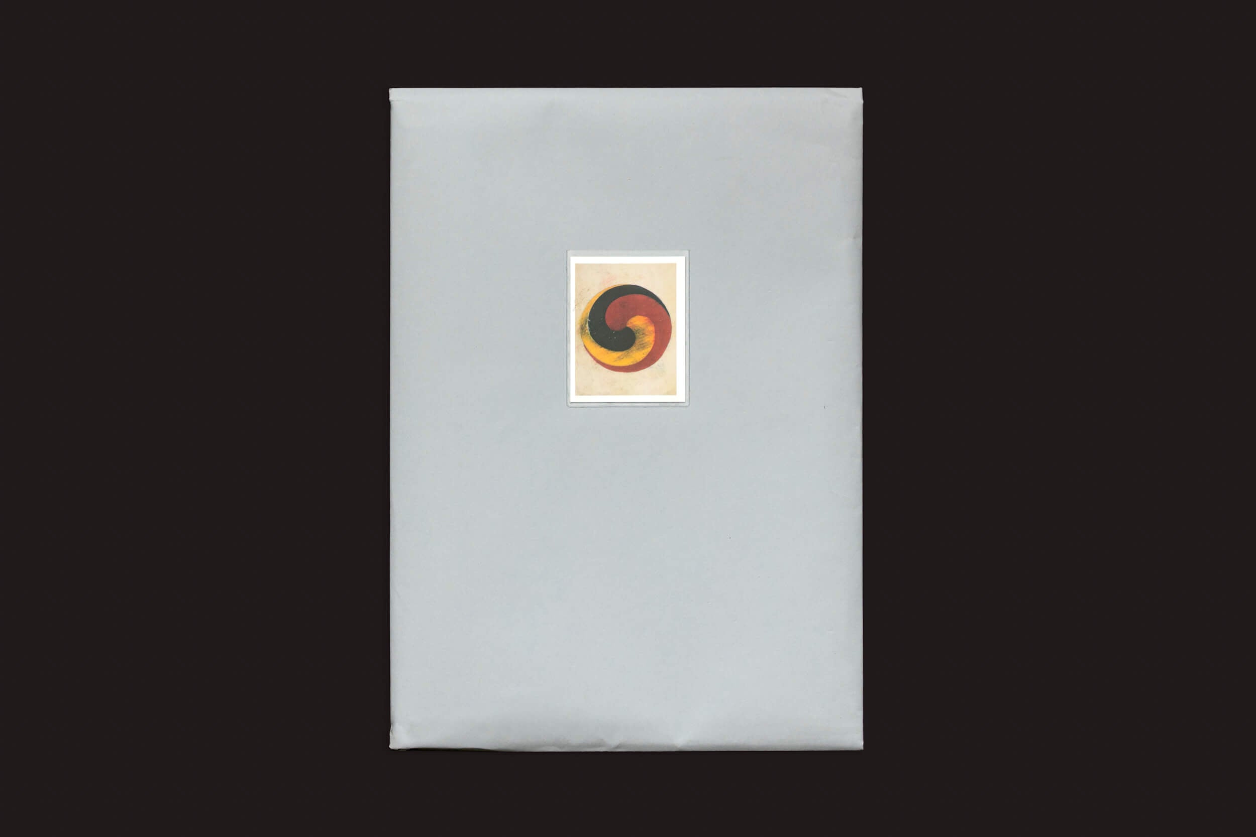 Um item retangular embrulhado em papel cinza decorado com um desenho circular abstrato representando Fruto, com tons de vermelho, laranja e preto em um fundo escuro, de Eduard Sánchez Ribot, da Handshake: Tierra, Camino, Fruto.