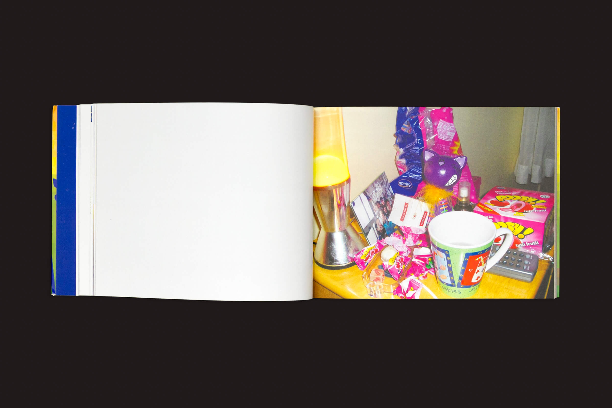 Um livro aberto, Amanda Devulsky: Ultrasuperfície da zero-Edições, revela uma foto colorida na página direita apresentando uma mesa com uma luminária de lava, uma caneca grande e uma embalagem de doces. A página esquerda está em branco, convidando pensamentos como um livro de artista no Fotolog.net pode capturar.