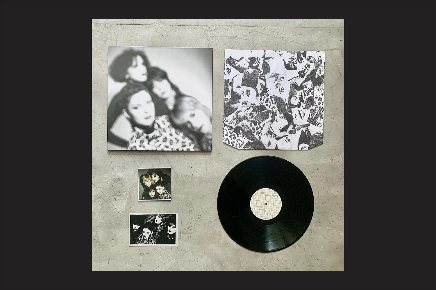 O vinil AKT 3: Frauen-Feuer em vinil da Nada Nada Discos, com capa do álbum em preto e branco, capa interna impressa combinando e dois pequenos cartões com fotos do supergrupo de quatro membros, são colocados em uma superfície cinza claro.