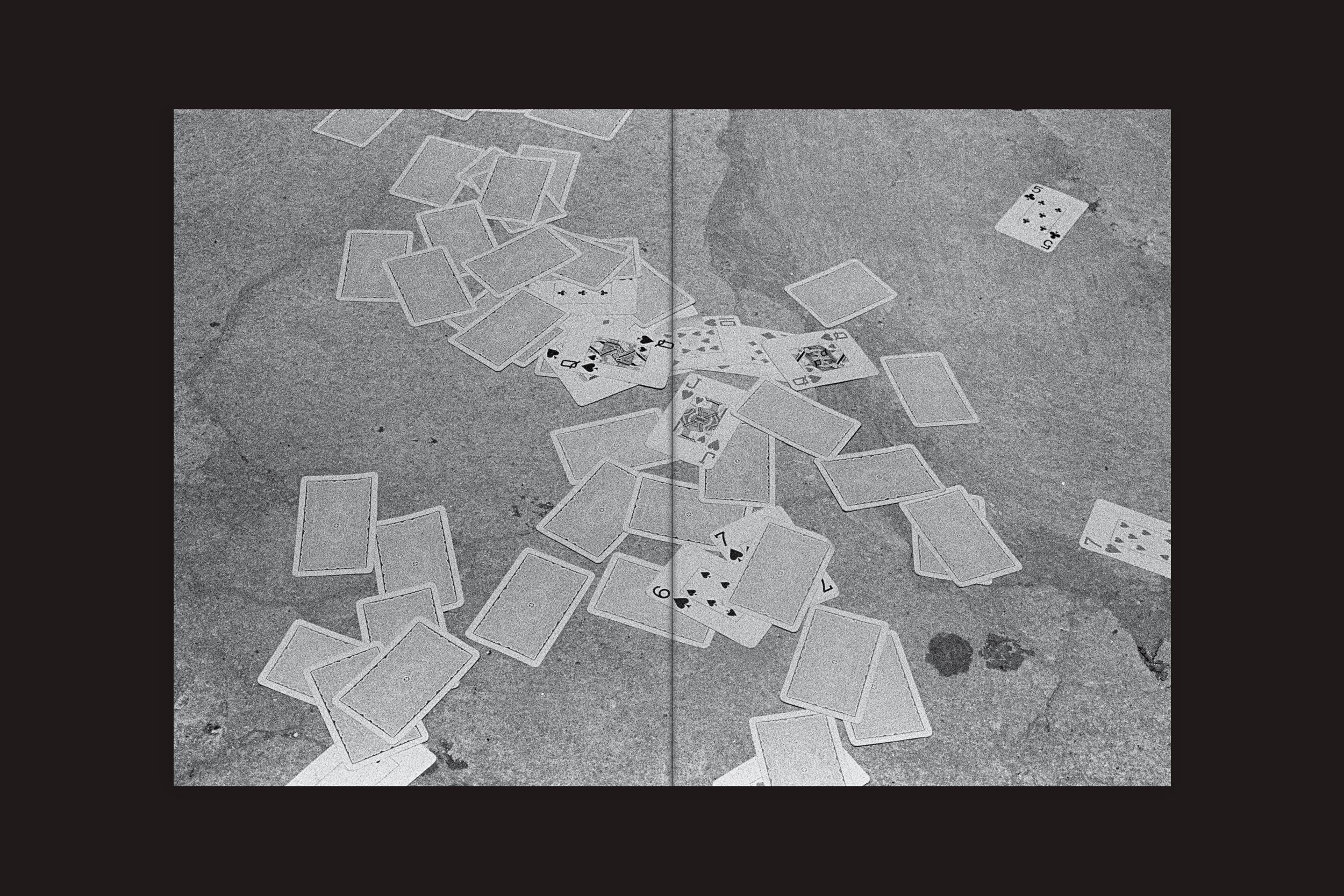 Uma pilha de cartas de baralho espalhadas sobre o concreto rachado, evocando a solidão. Essa imagem em preto e branco enquadra a existência fragmentada, conforme apresentado em "André Djanikian: Algunos sentimientos no cambian junto a las estaciones", de Selo Turvo.