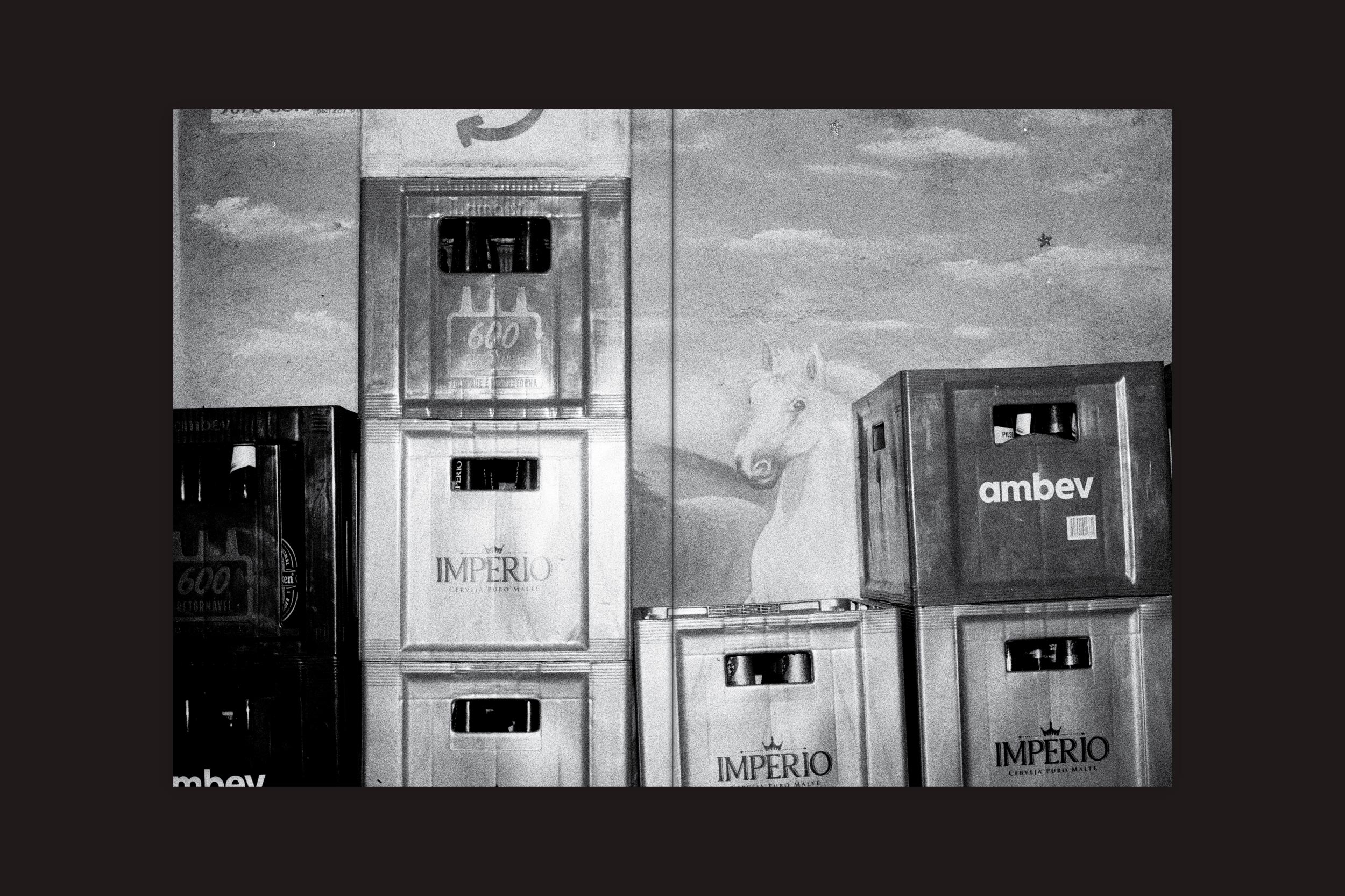 Pilhas de caixas vazias estão diante de um mural de um cavalo branco sob um céu nublado, evocando solidão. A foto, em preto e branco, lembra a memória de André Djanikian: Algunos sentimientos no cambian junto a las estaciones de Selo Turvo.