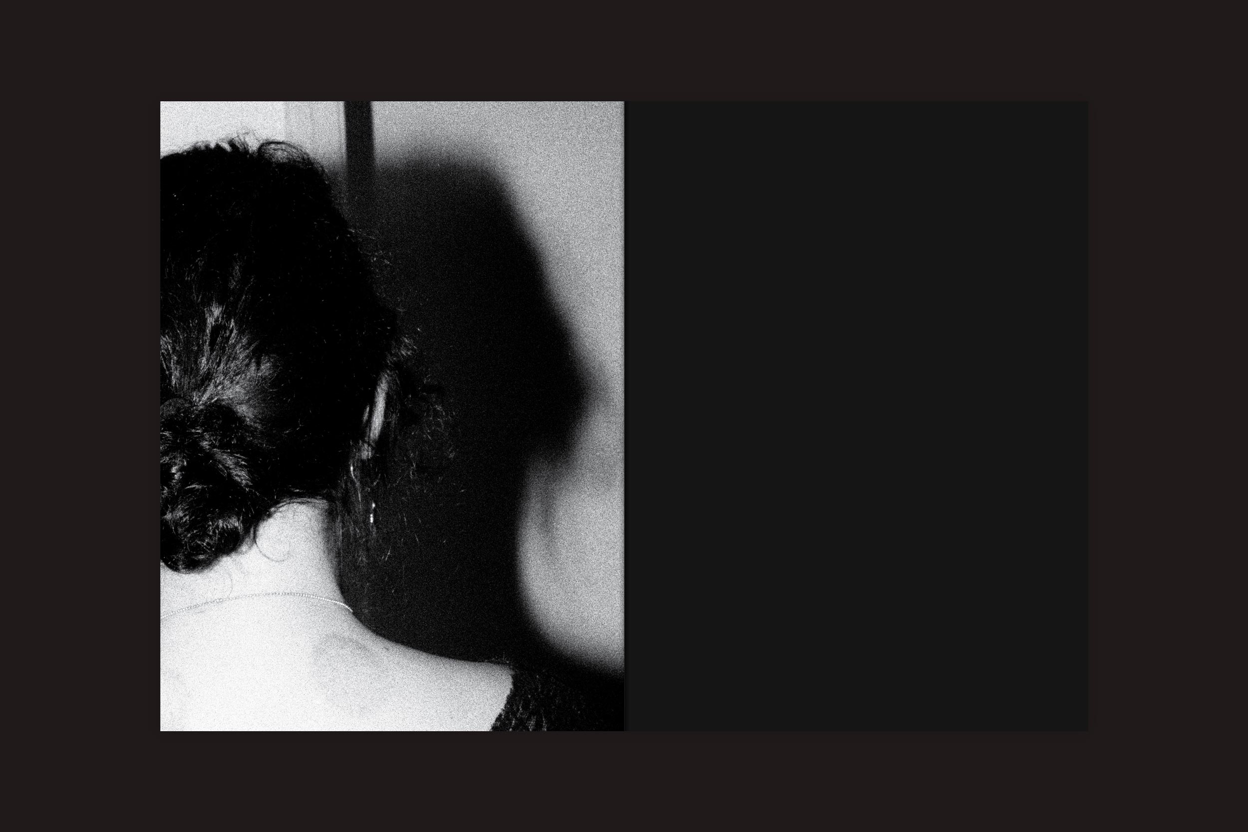 Uma mulher de cabelos escuros está de costas, com sua sombra projetada em uma parede predominantemente preta. Essa foto em preto e branco para a exposição "André Djanikian: Algunos sentimientos no cambian junto a las estaciones", do Selo Turvo, evoca solidão e mistério.